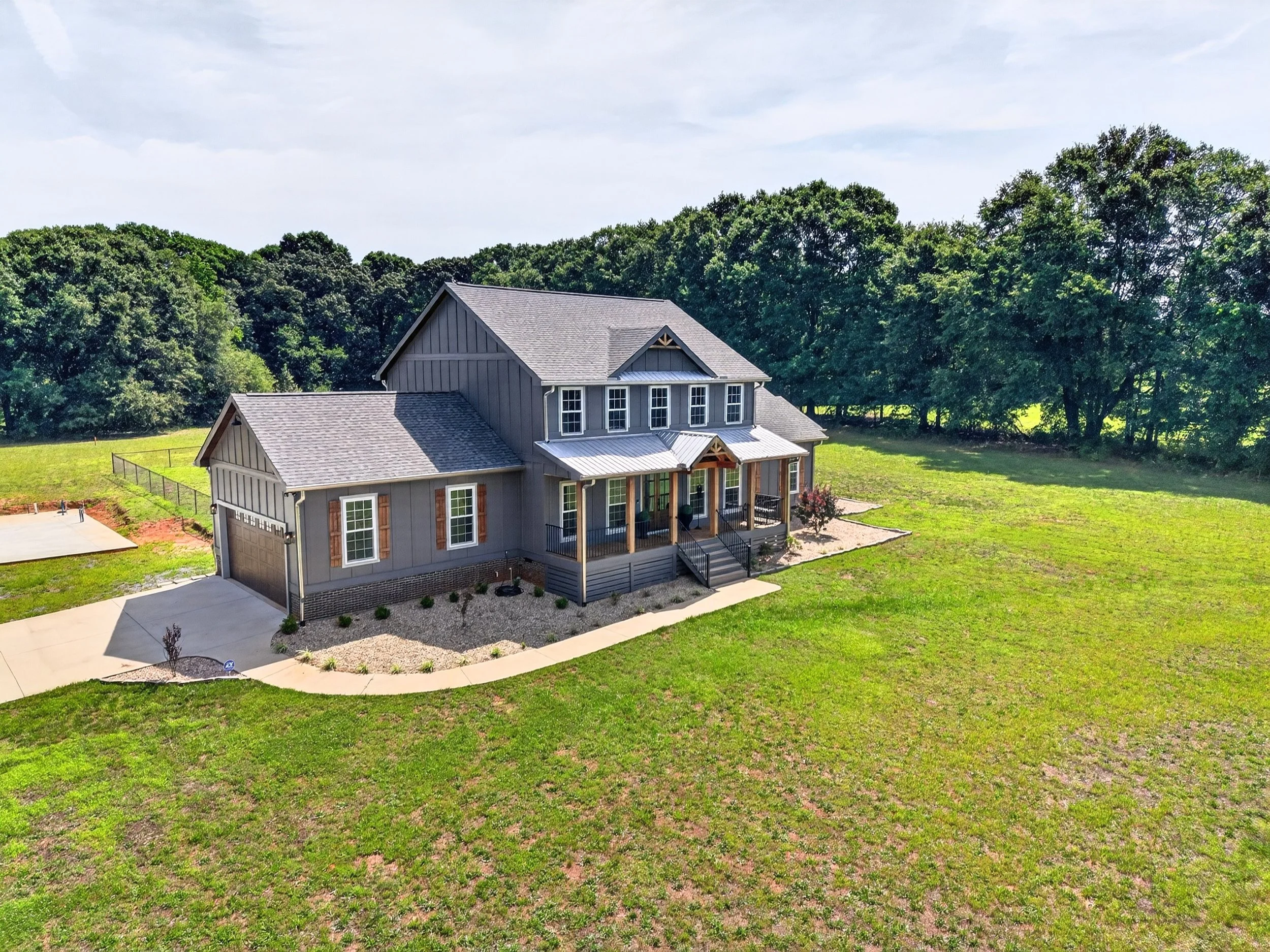 Large house with gray siding, multiple windows, a covered front porch, surrounded by a green lawn and trees in the background.
