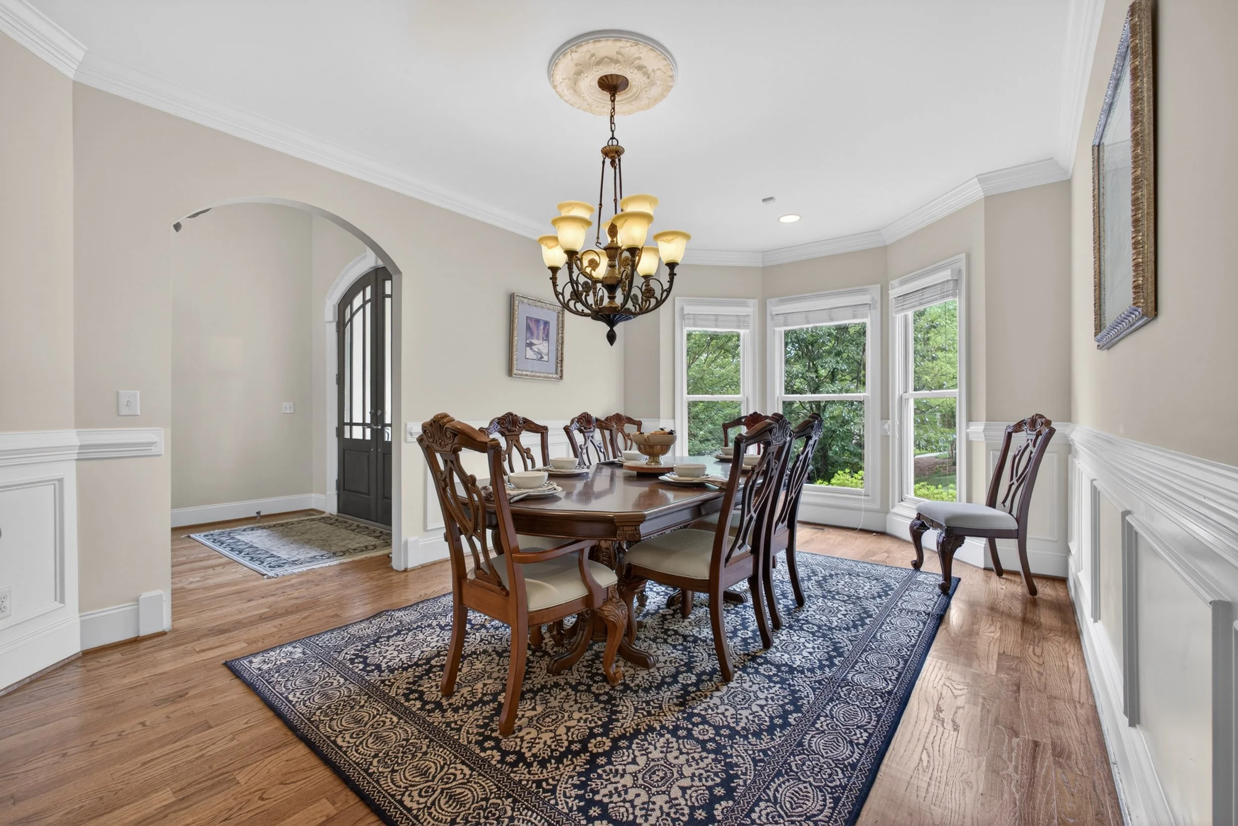 A dining room with a large wooden table, ten carved wooden chairs, a blue and beige patterned rug, a chandelier, three large windows, and artwork on the walls, with hardwood floors and beige walls.