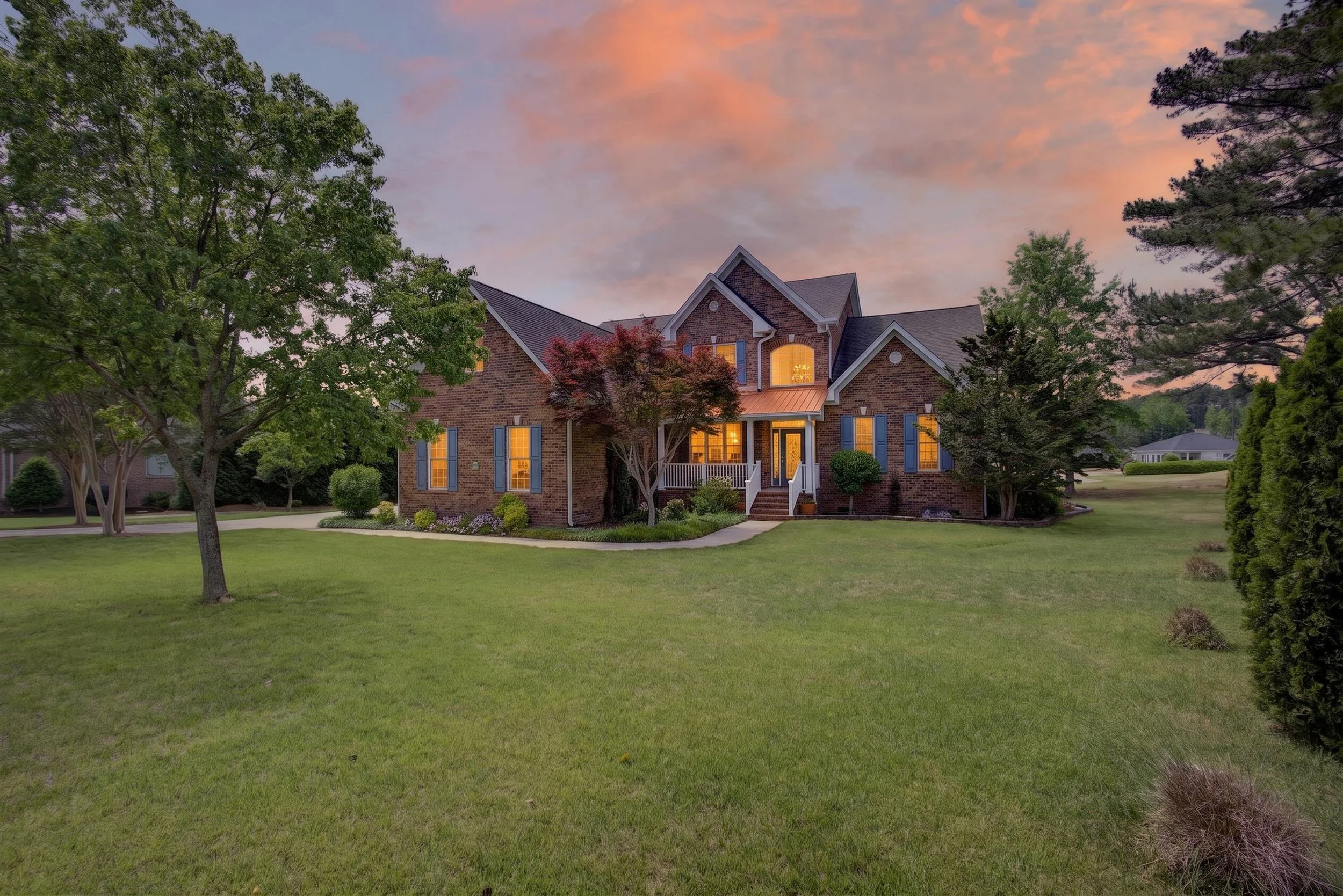 A brick house with lit windows at sunset, surrounded by green trees and a well-maintained lawn.