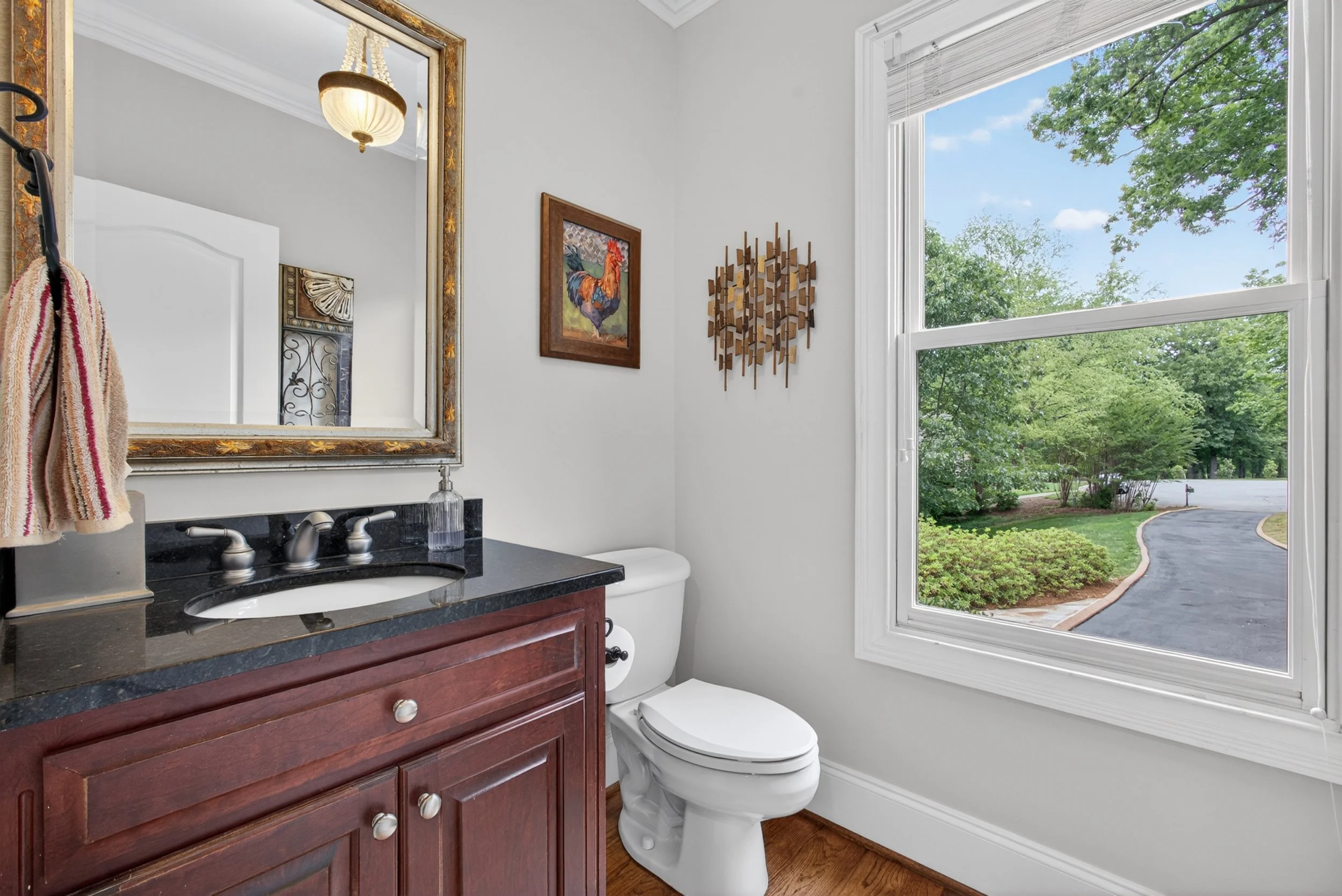 A bathroom with a large window showing trees and a driveway outside, a mirror above the sink, artwork on the wall, a countertop with a sink, and a toilet next to the window.
