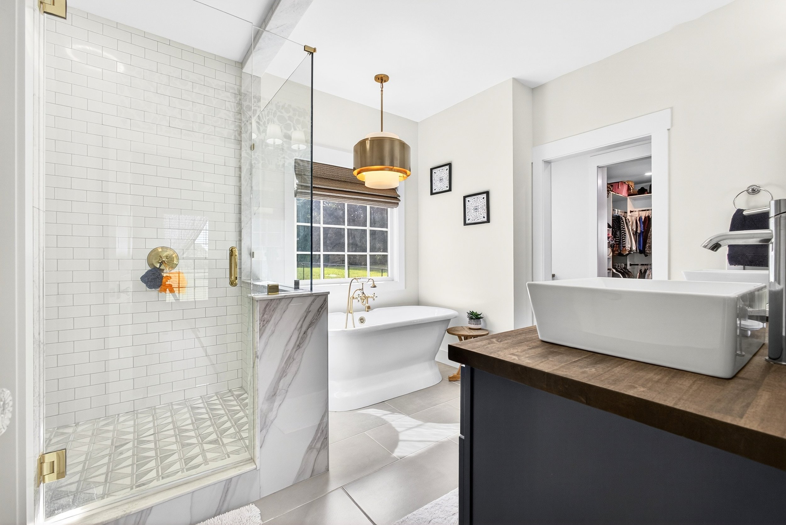 Modern bathroom with a glass-enclosed shower, a clawfoot bathtub, a wooden vanity with a vessel sink, a large window, and walk-in closet visible through an open door.
