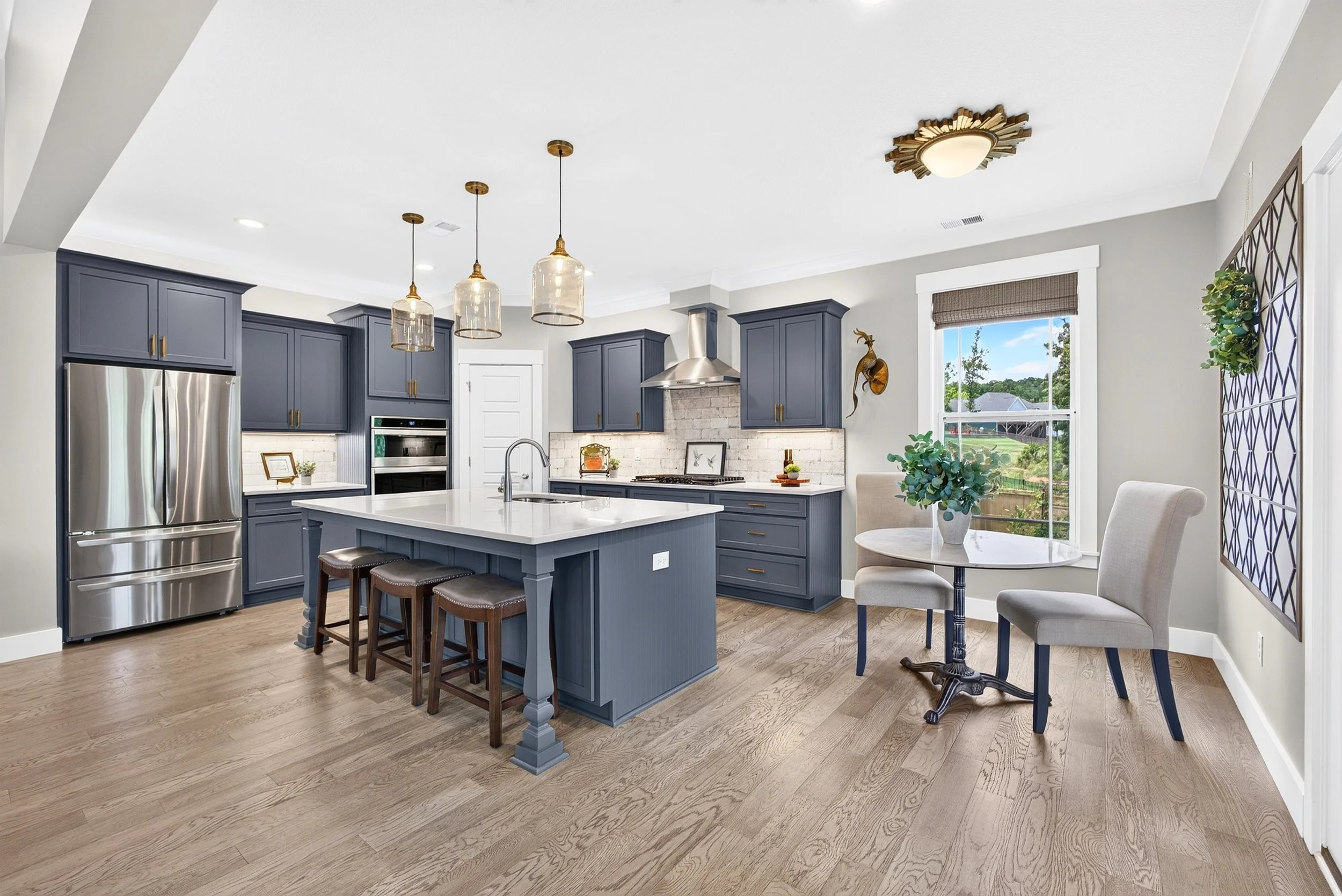 Modern kitchen with navy blue cabinets, stainless steel appliances, white marble island, and wooden flooring. There are three pendant lights above the island, a window with a view outside, and a small dining table with chairs.