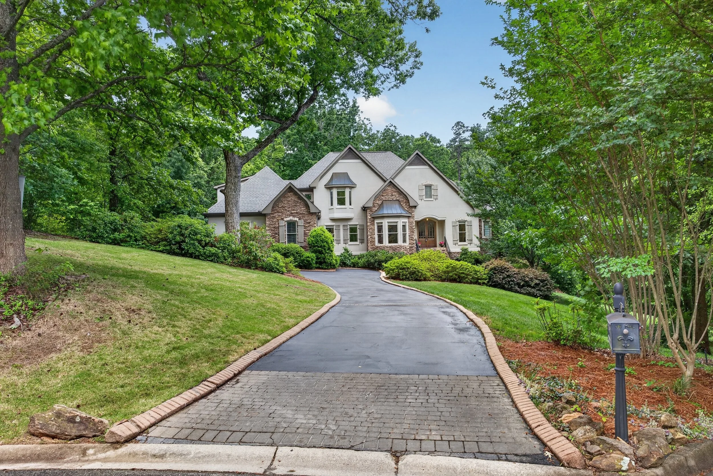A paved driveway leading to a large house surrounded by greenery, trees, and shrubs, with a mailbox at the front.