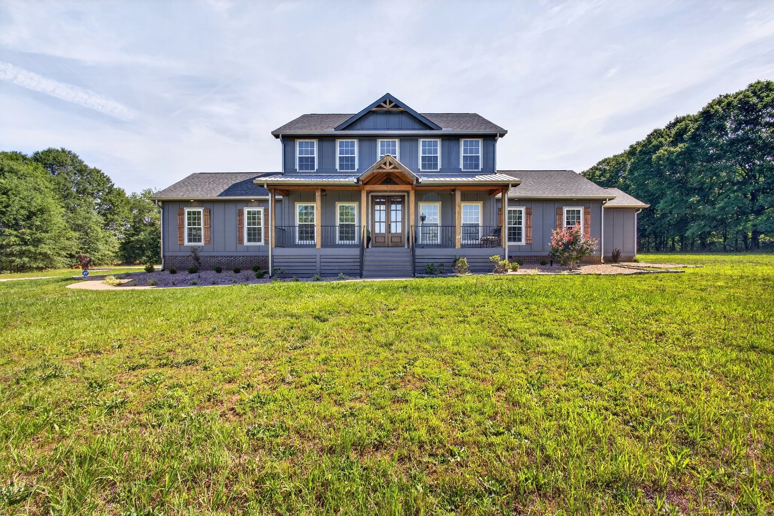 Front view of a two-story blue house with a front porch, surrounded by a grassy yard and trees under a partly cloudy sky.