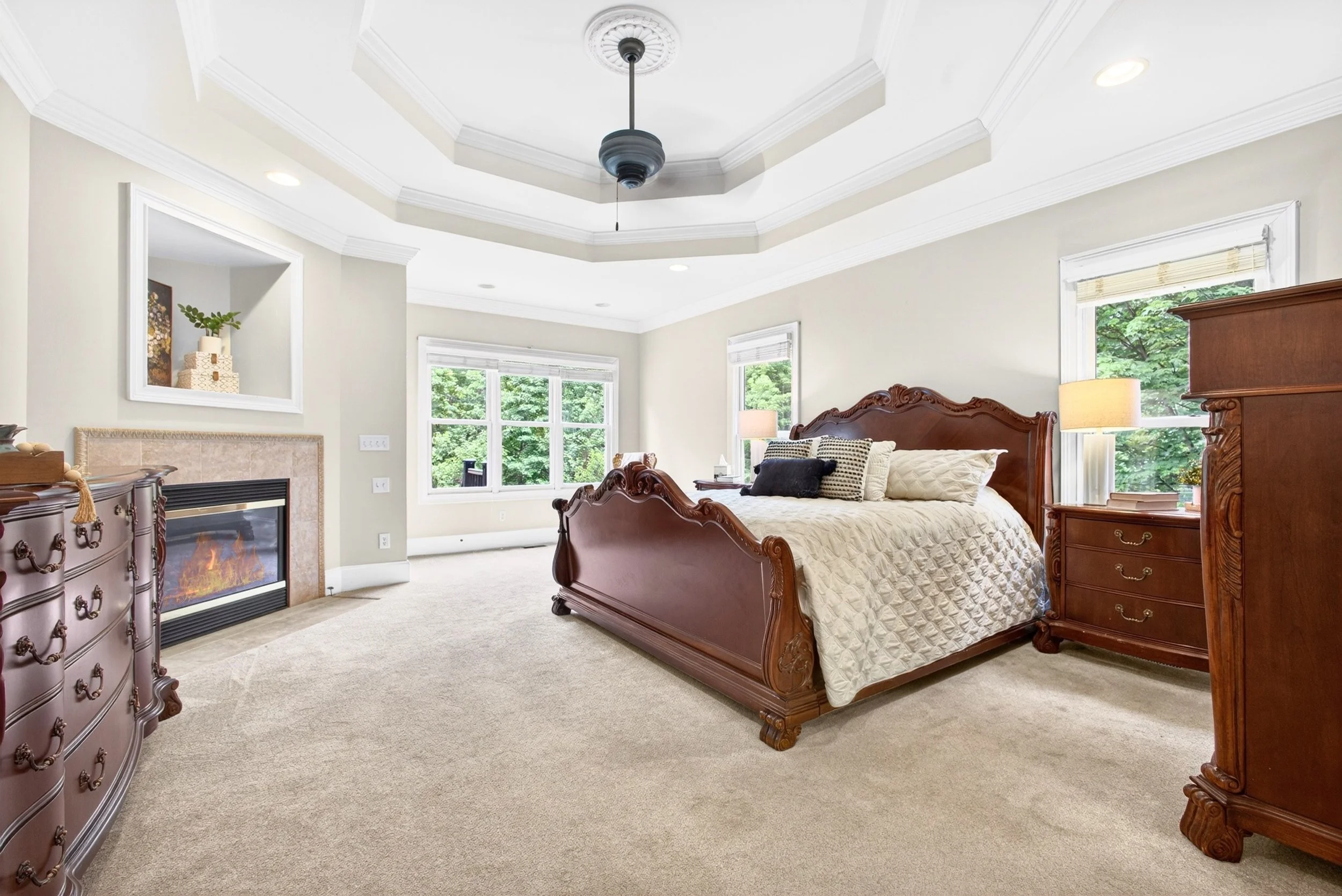 A bedroom with a wooden bed, large windows showing green trees outside, a fireplace, and a TV on a dresser.
