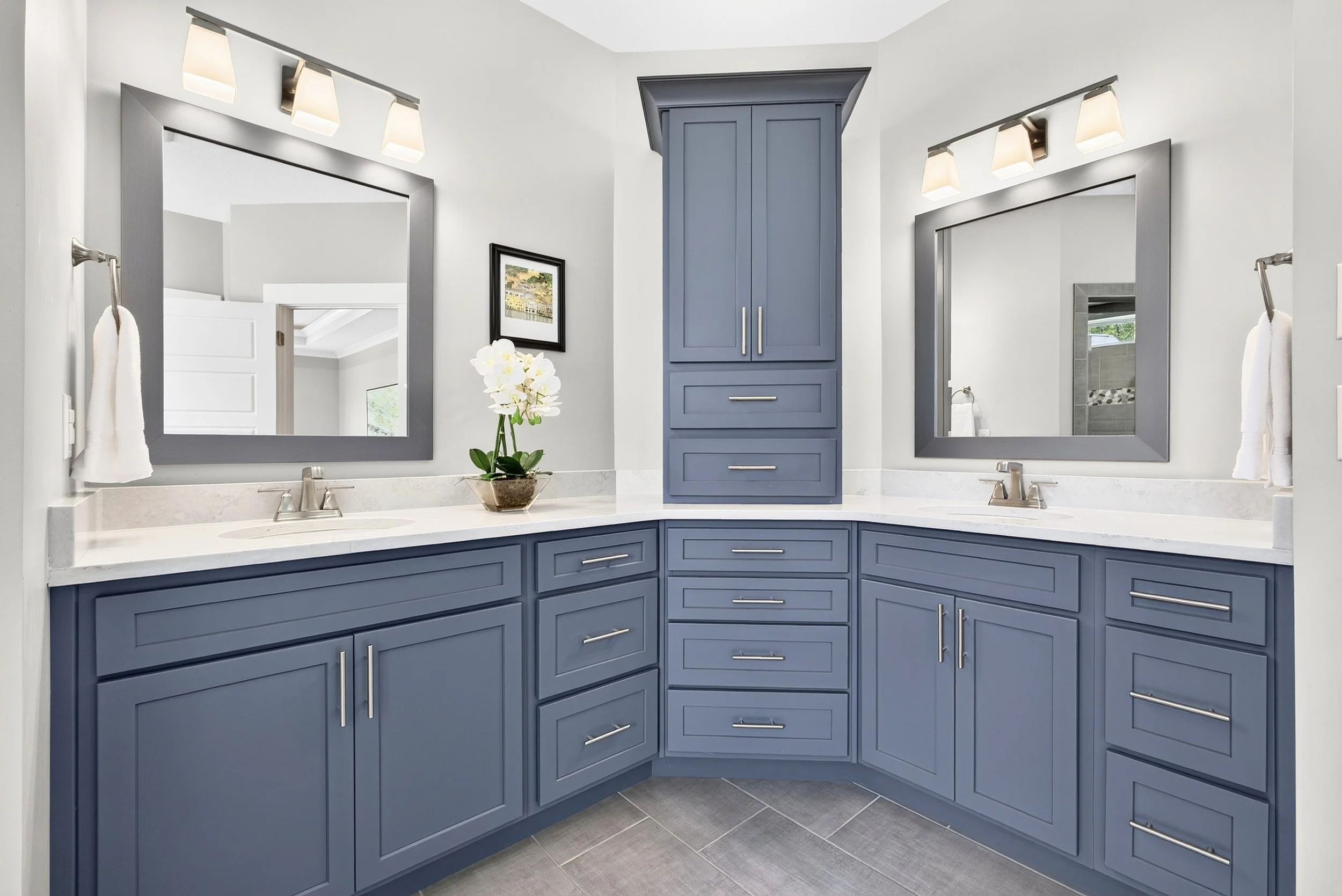Bathroom with blue cabinets, two large mirrors, white countertops, and a plant with white flowers on the counter.