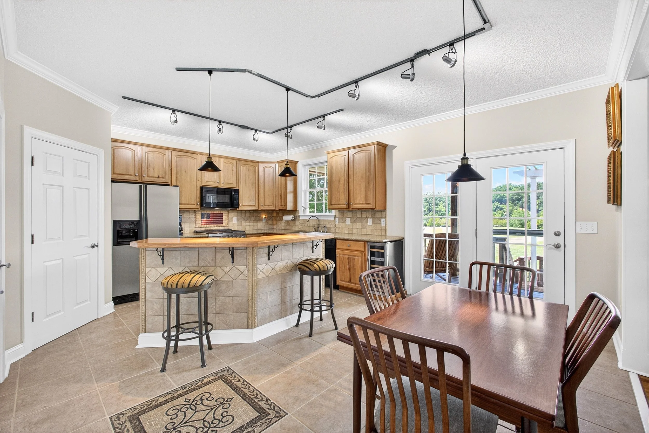 Kitchen with wooden cabinets, stainless steel refrigerator, microwave, tile backsplash, and pendant lights hanging over a breakfast bar with three stools. Large window and glass door lead outside, with a view of greenery.