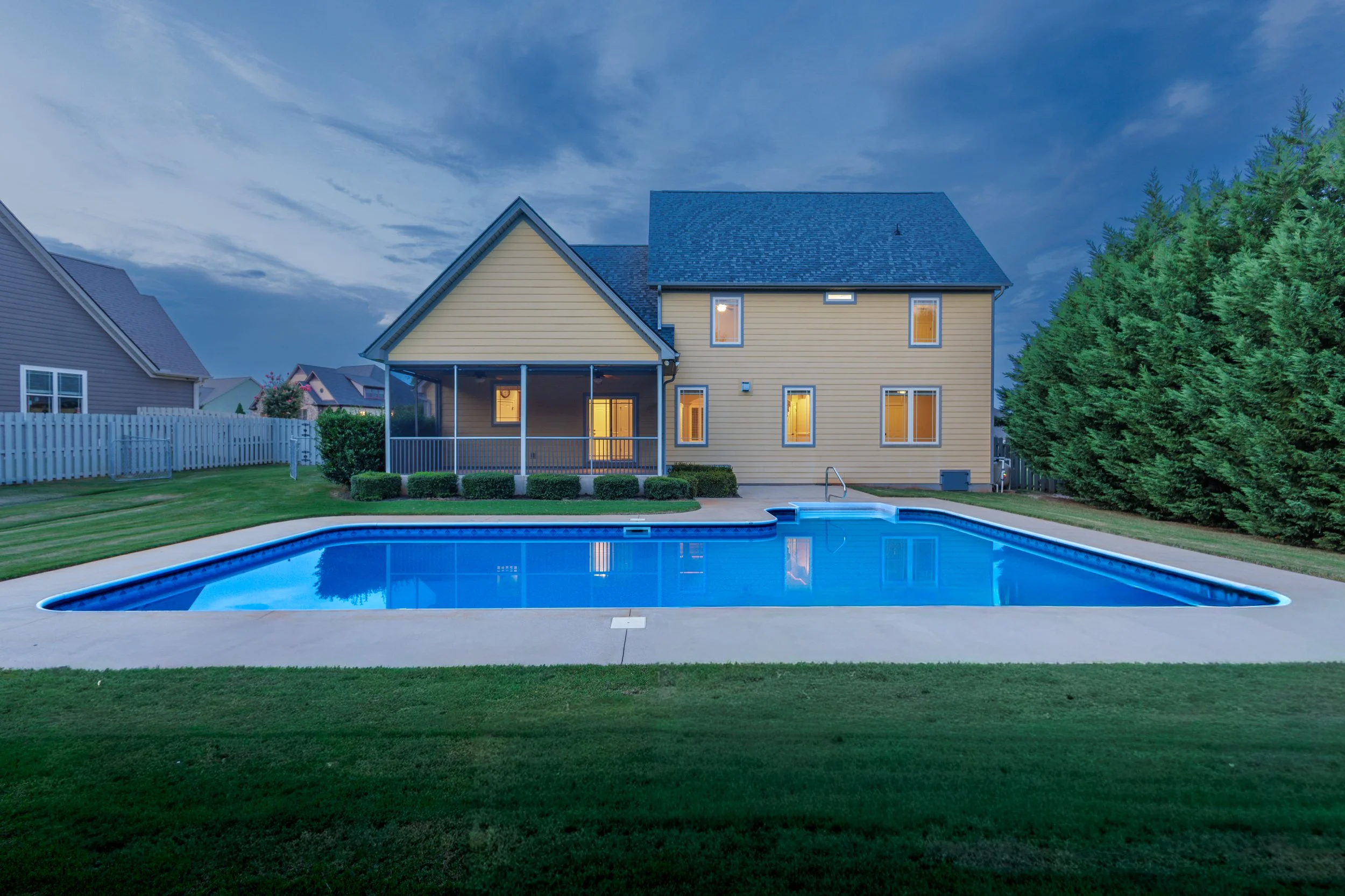 Backyard with swimming pool, green lawn, large house with beige siding, multiple windows, and a screened porch, during evening with lights on inside the house, surrounded by neighboring houses and trees.