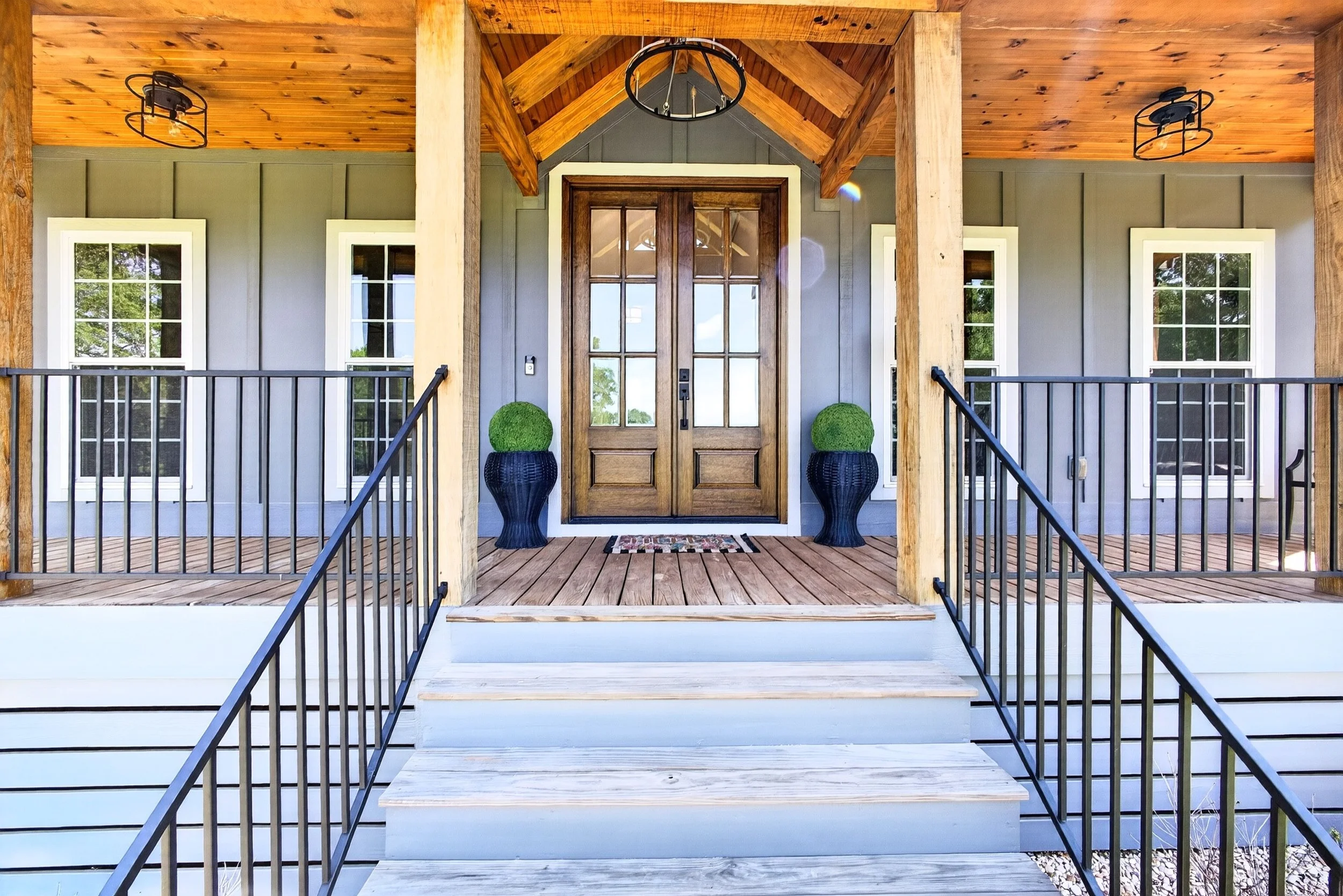 Front porch of a house with a wooden door, black railing, and potted topiary plants, with a wooden ceiling and beams.
