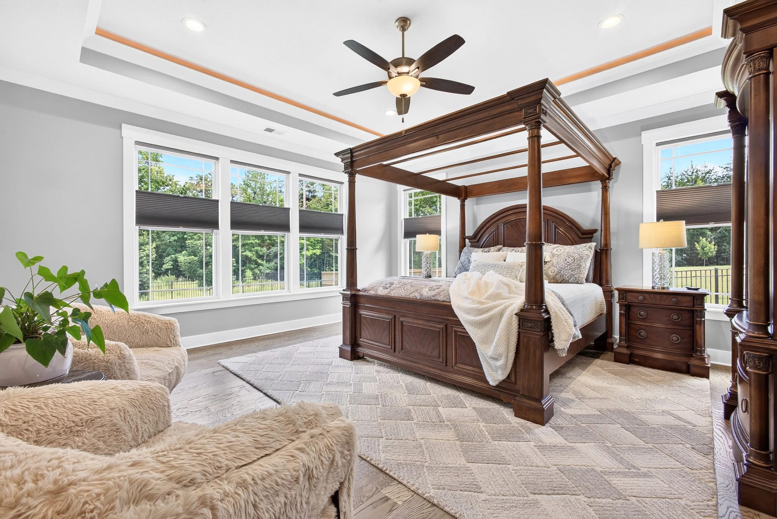 A bedroom with a wooden four-poster bed, two nightstands with lamps, large windows, a cozy beige chair, and a ceiling fan.
