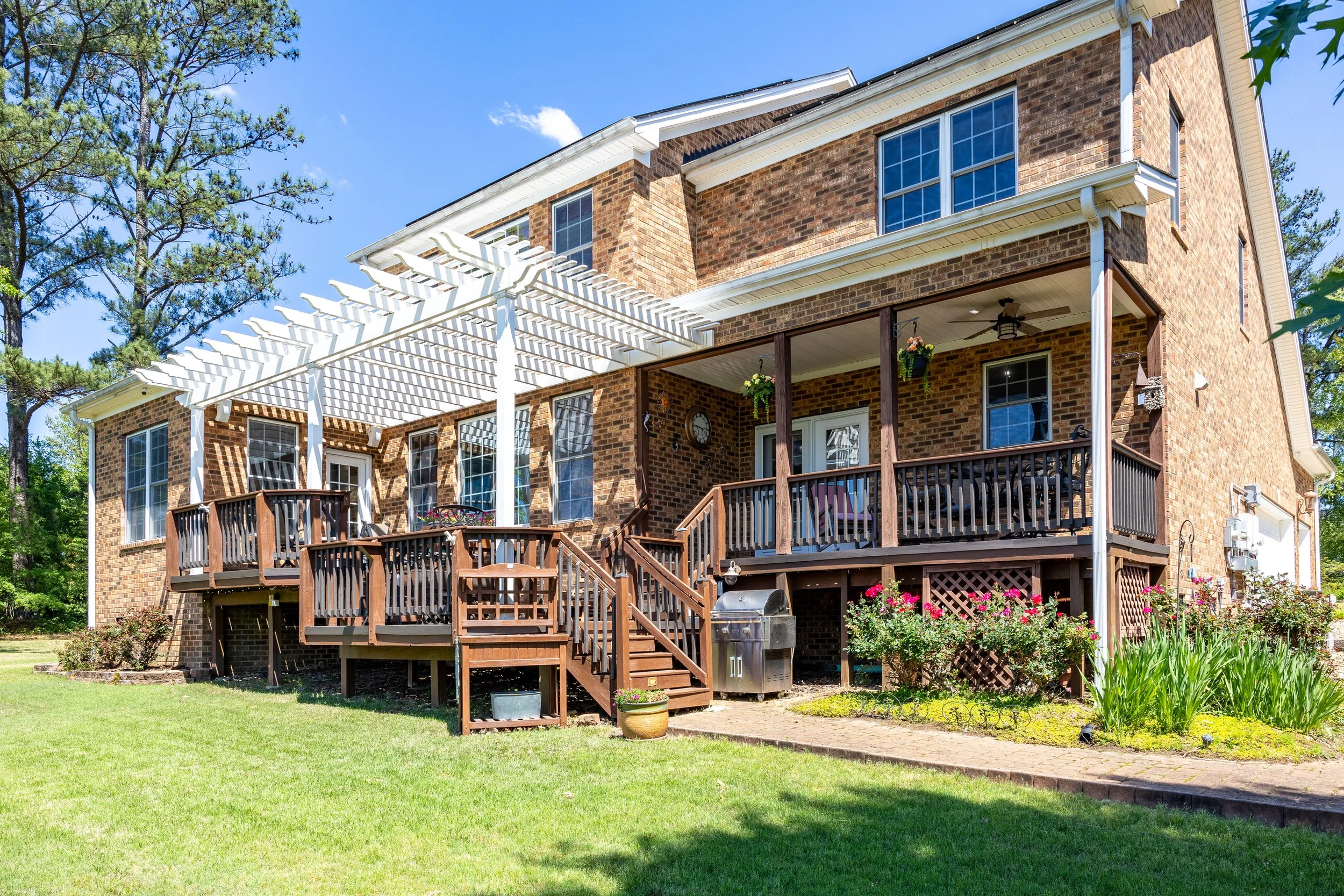 A two-story brick house with a wooden deck and railing, potted plants, hanging baskets, a ceiling fan, and an outdoor grill, surrounded by a green lawn and trees under a blue sky.