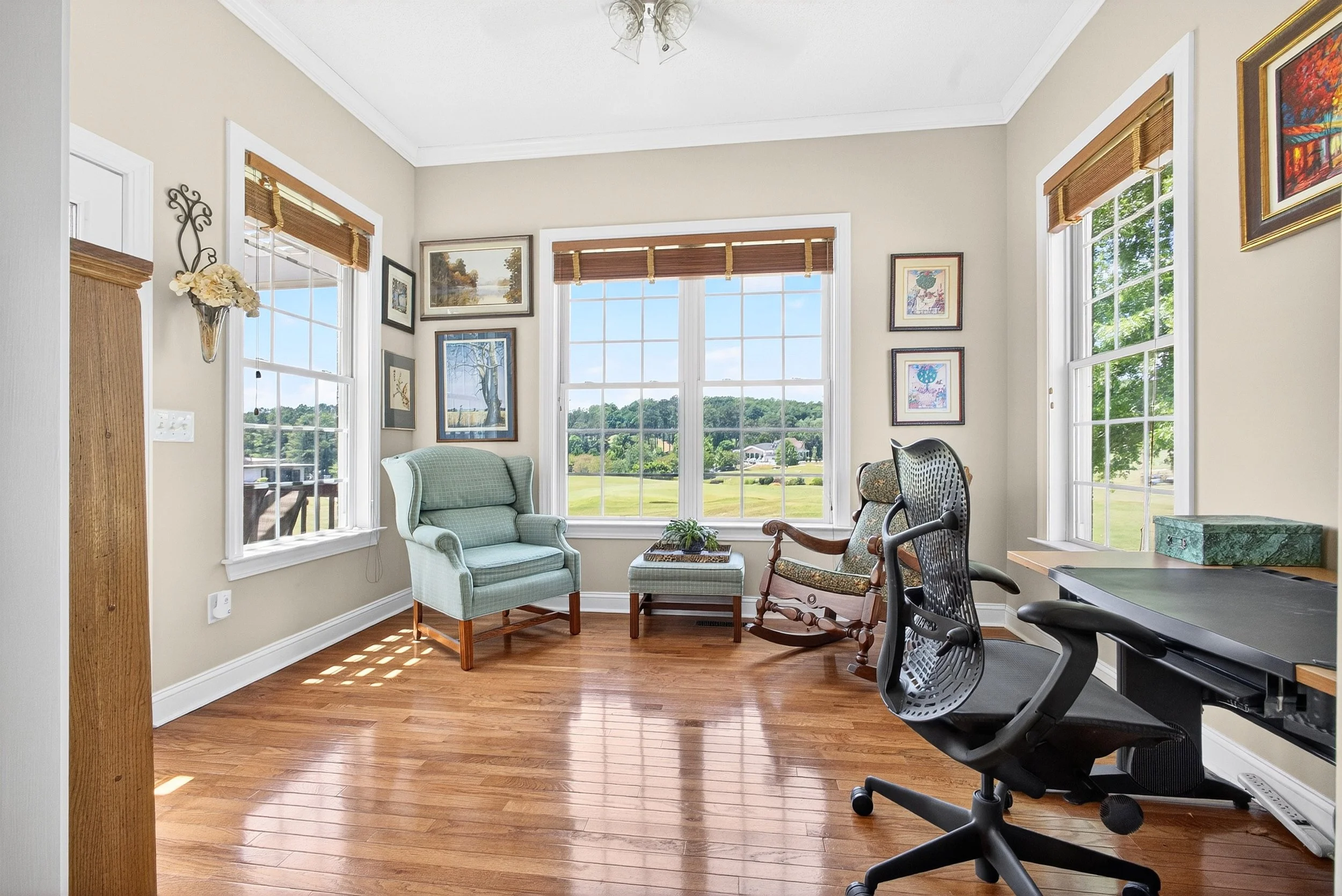 The image shows a bright, cozy room with large windows revealing a green landscape outside. The interior includes a seating area with two upholstered chairs, one in light green and the other with patterned fabric, a small table with a plant, and some