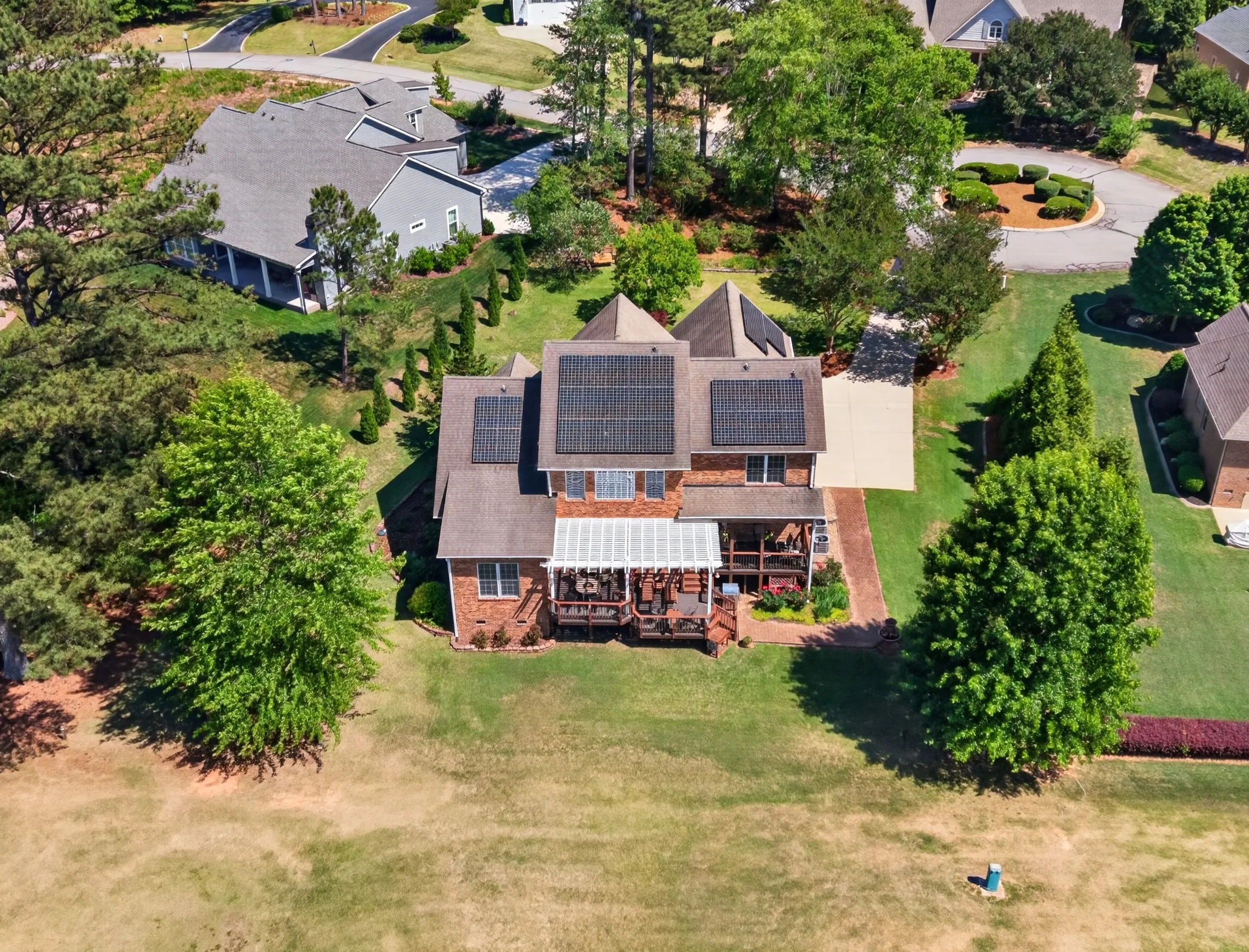 An aerial view of a residential neighborhood with multiple houses, yards, and trees. One house has solar panels on the roof, and there's a small lawn area with a fire pit. Paved roads surround the houses with some curving, and there is a patch of yel