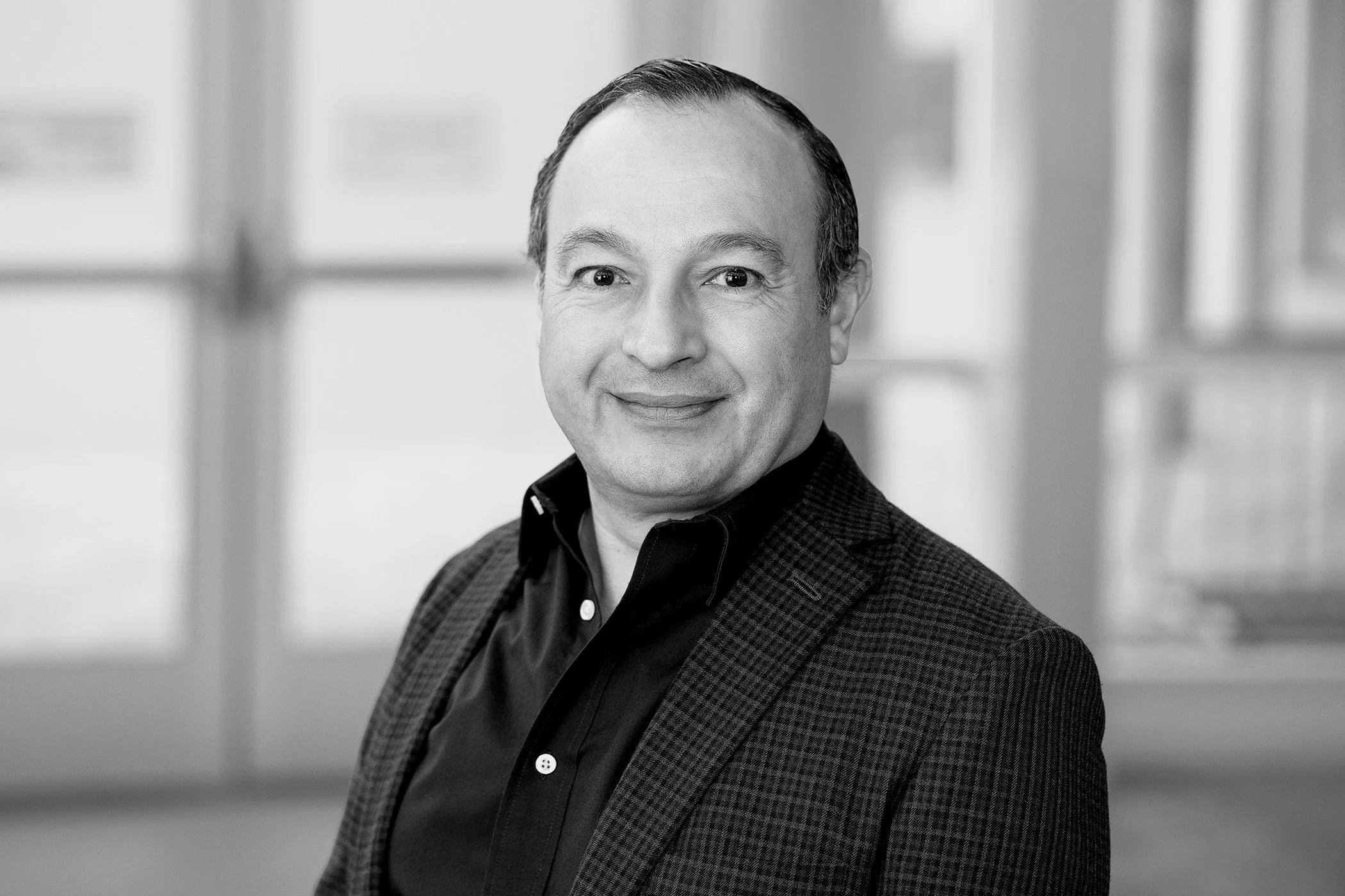 Black and white portrait of a middle-aged man with short hair, wearing a checkered blazer and dark shirt, smiling gently. He is indoors with blurred background suggesting a professional setting.