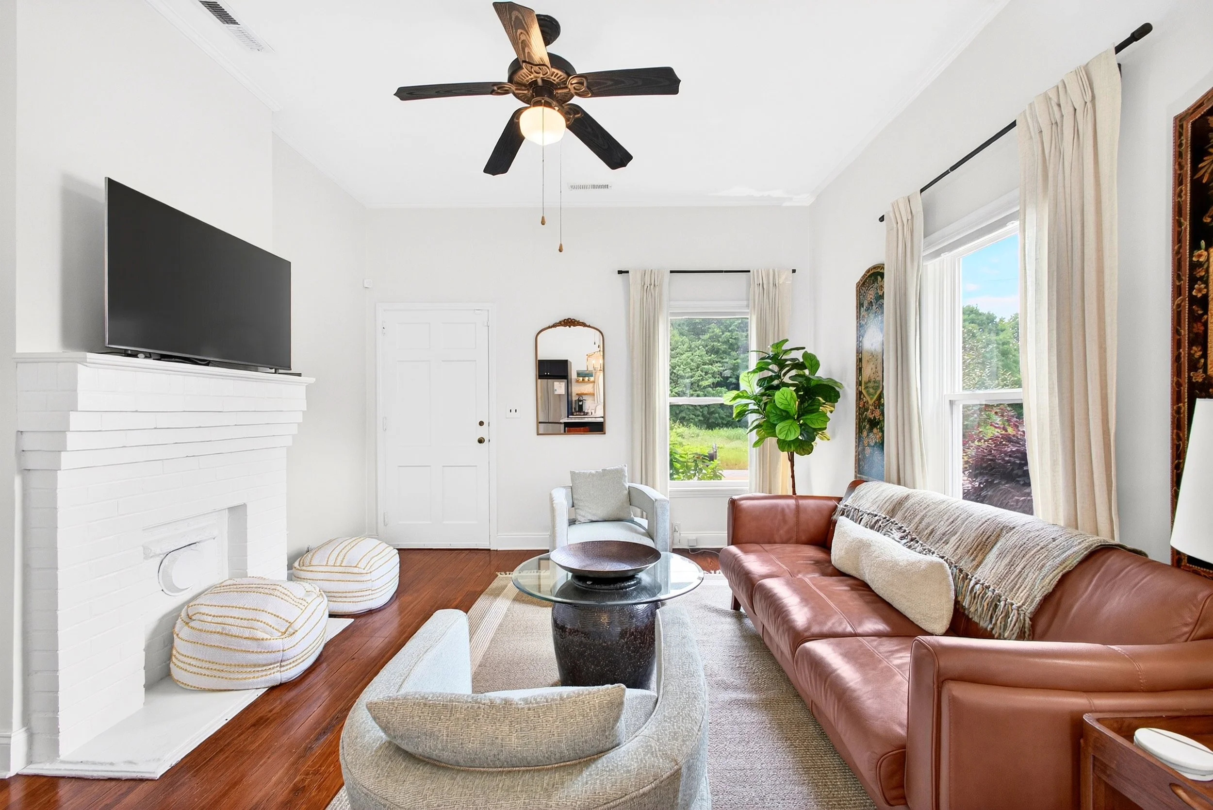 Living room with a white brick fireplace, a wall-mounted flat-screen TV, large windows with cream curtains, a green potted plant, a brown leather sofa, a white armchair, round ottomans, a glass coffee table, and a ceiling fan with black blades and a light.
