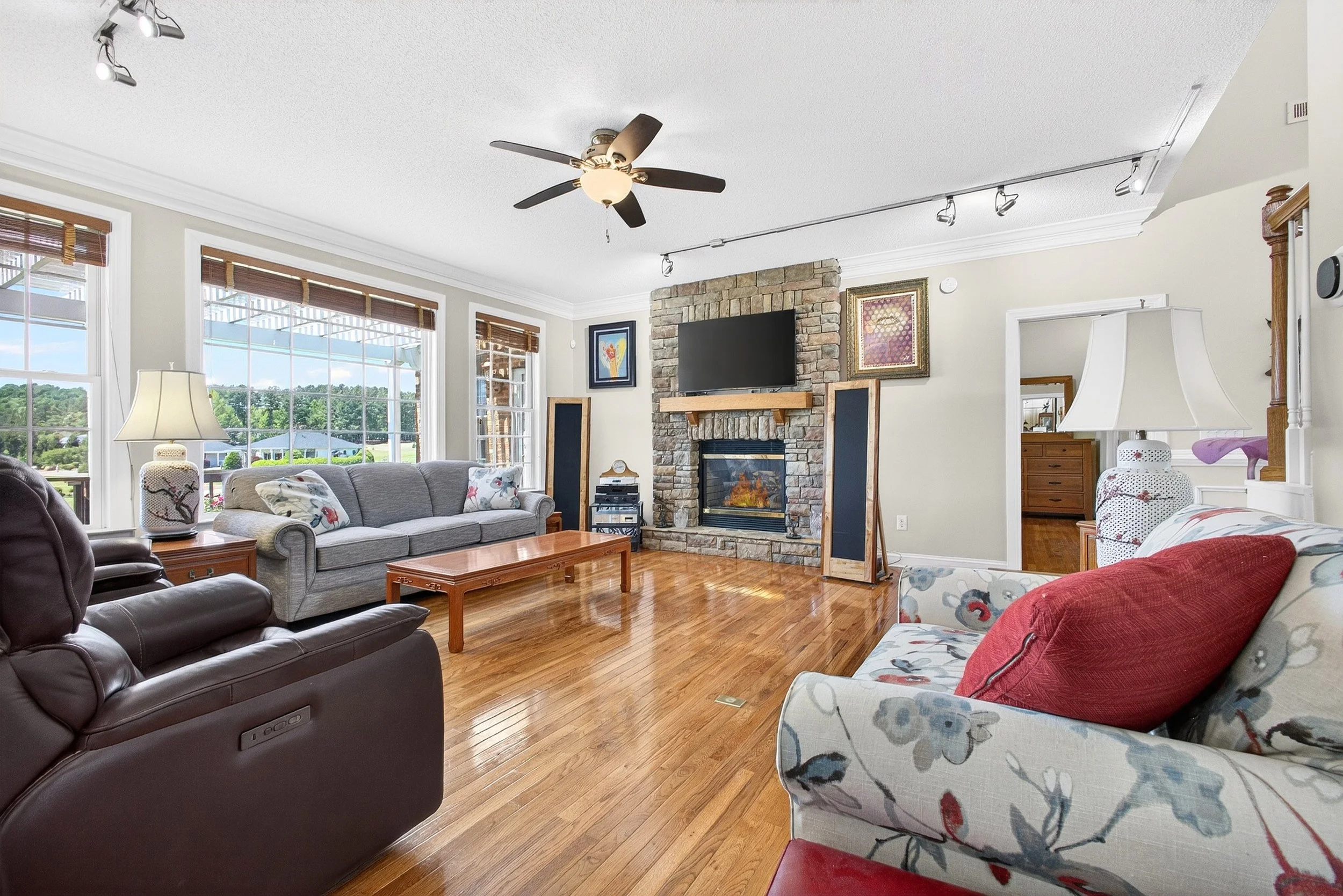 Living room with large windows, hardwood floors, a stone fireplace, various seating including a brown recliner, floral upholstered armchair, and a grey sofa, wooden coffee table, lamps, artwork on the walls, and a ceiling fan.