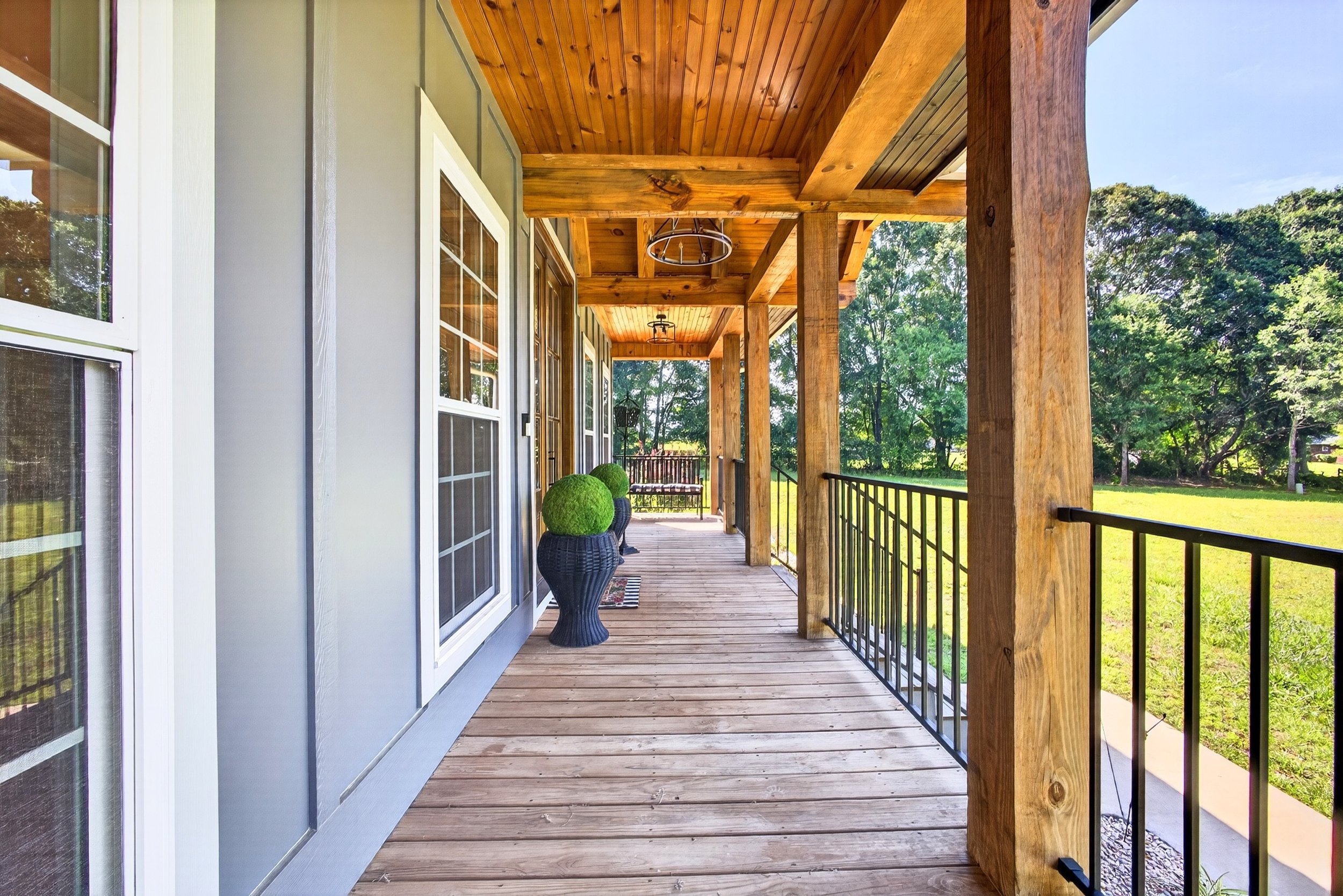 Wooden porch with railing and plants in pots, exterior house wall on the left, trees and a grassy yard in the background.