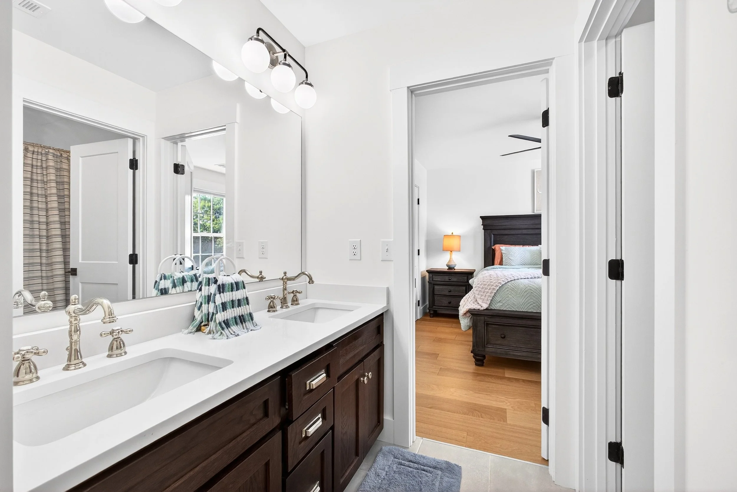 Bathroom with double vanity mirror, dark wooden cabinets, white countertop, silver faucets, towels, and a view into a bedroom with a bed, nightstand, and lamp.