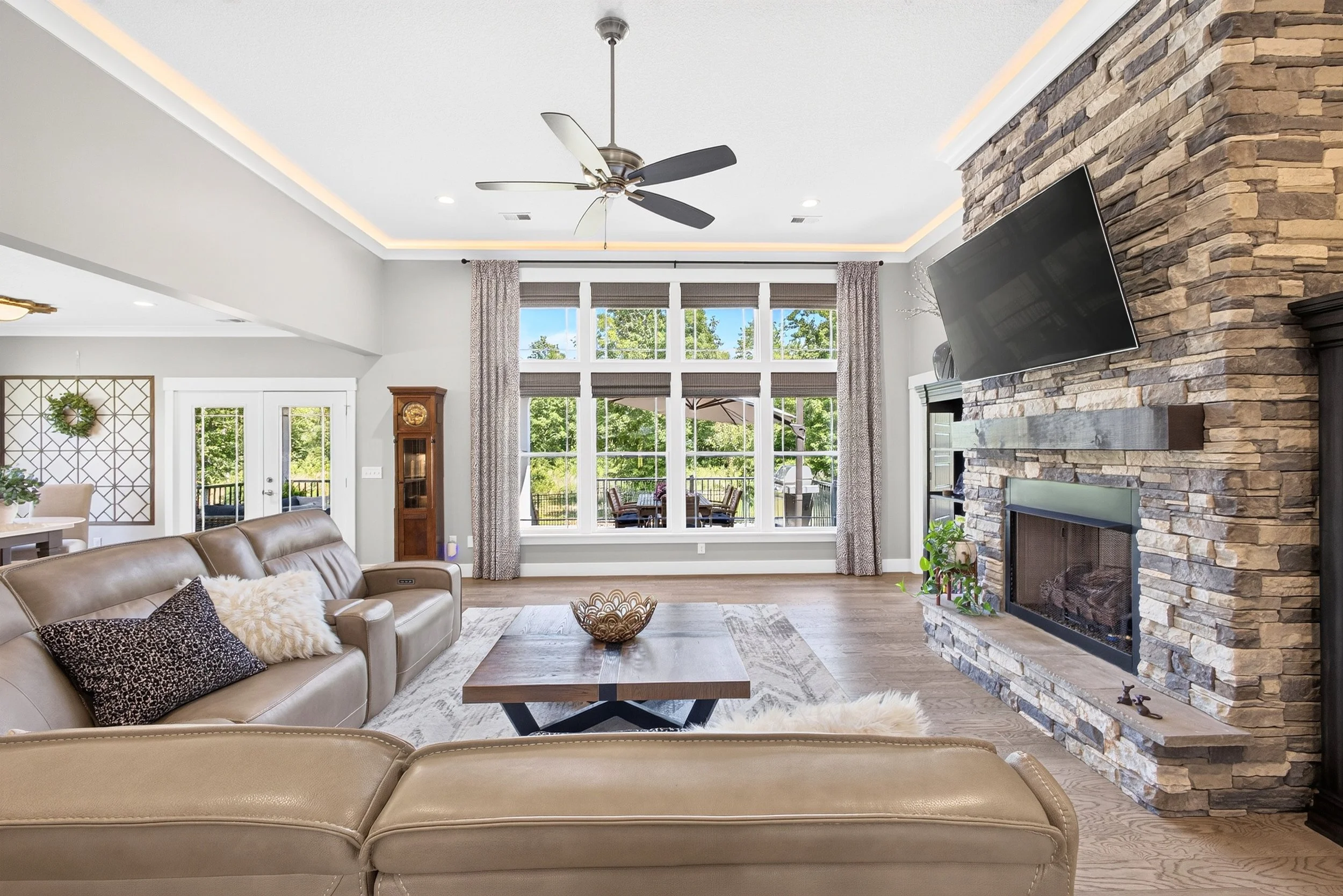 Living room with beige leather sofa, stone fireplace with mounted TV, large window with curtains, ceiling fan, and outdoor dining area visible through the window.