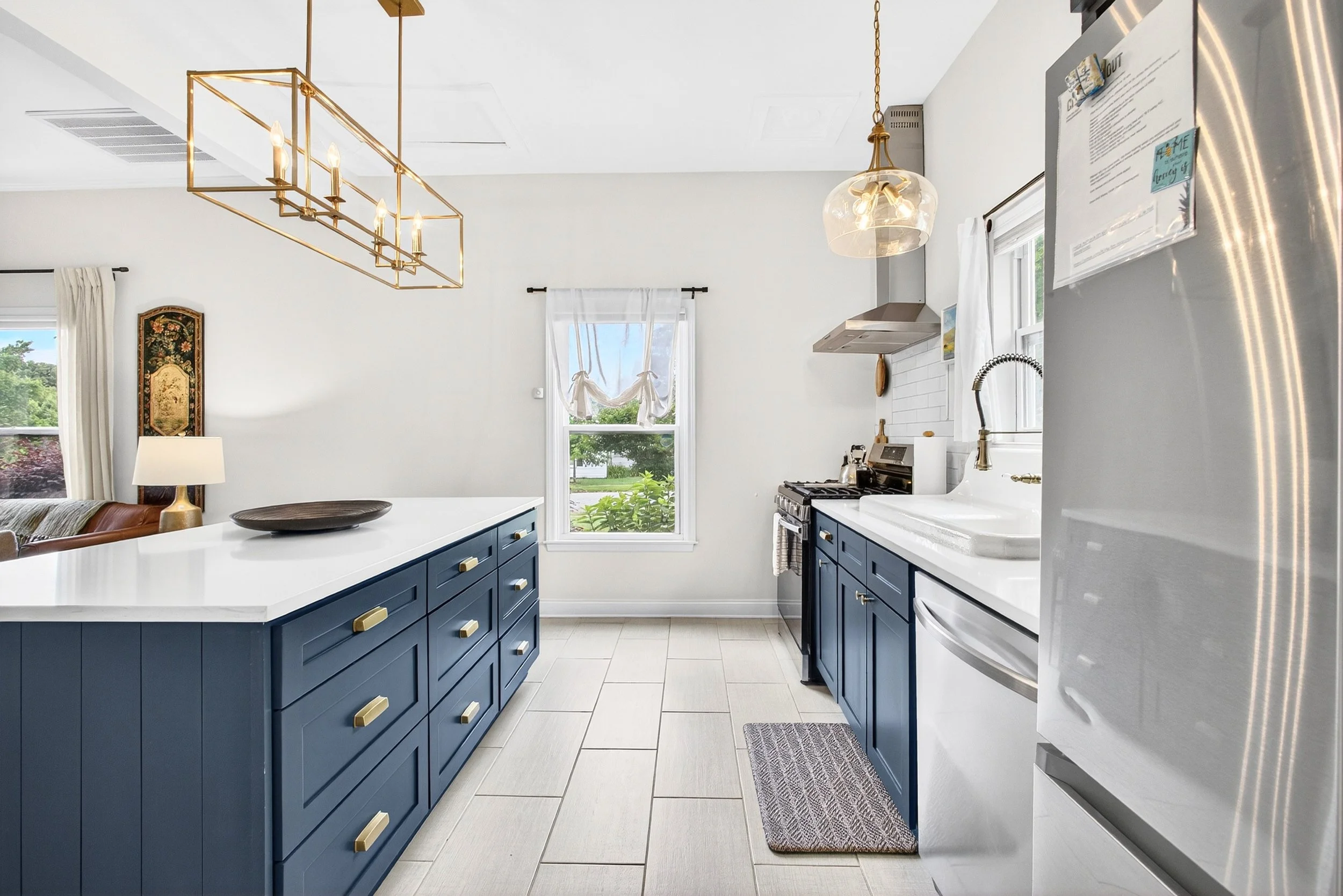A bright kitchen with white walls, blue cabinets with gold handles, a white countertop, stainless steel appliances, pendant lights, and large windows with white curtains showing outdoor greenery.