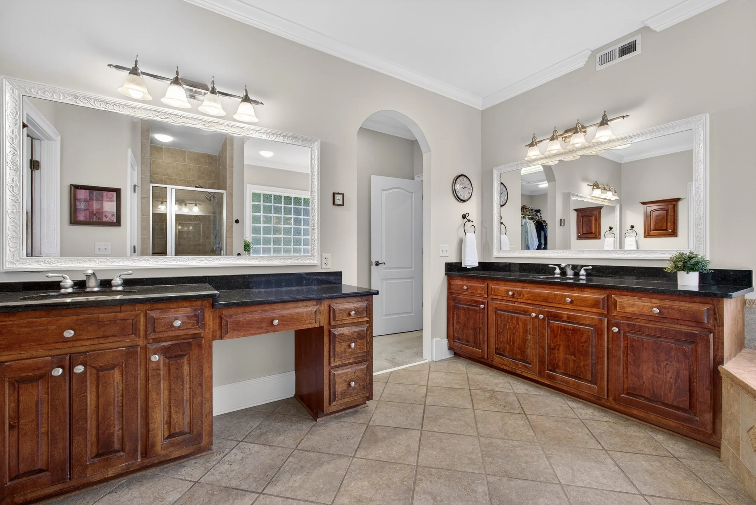 A bathroom with two large mirrors, wooden cabinets, black countertops, and dual sinks. The room has beige walls, tiled floors, and a walk-in shower with a glass door.