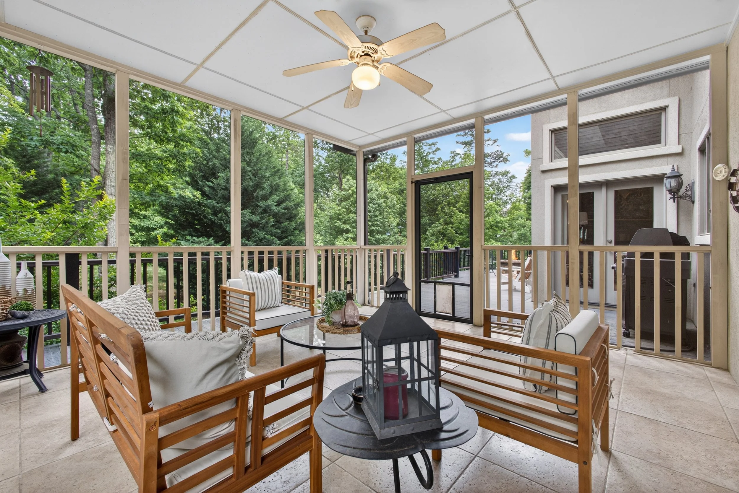 Covered porch with wooden furniture, pillows, decorative lantern, and greenery view.