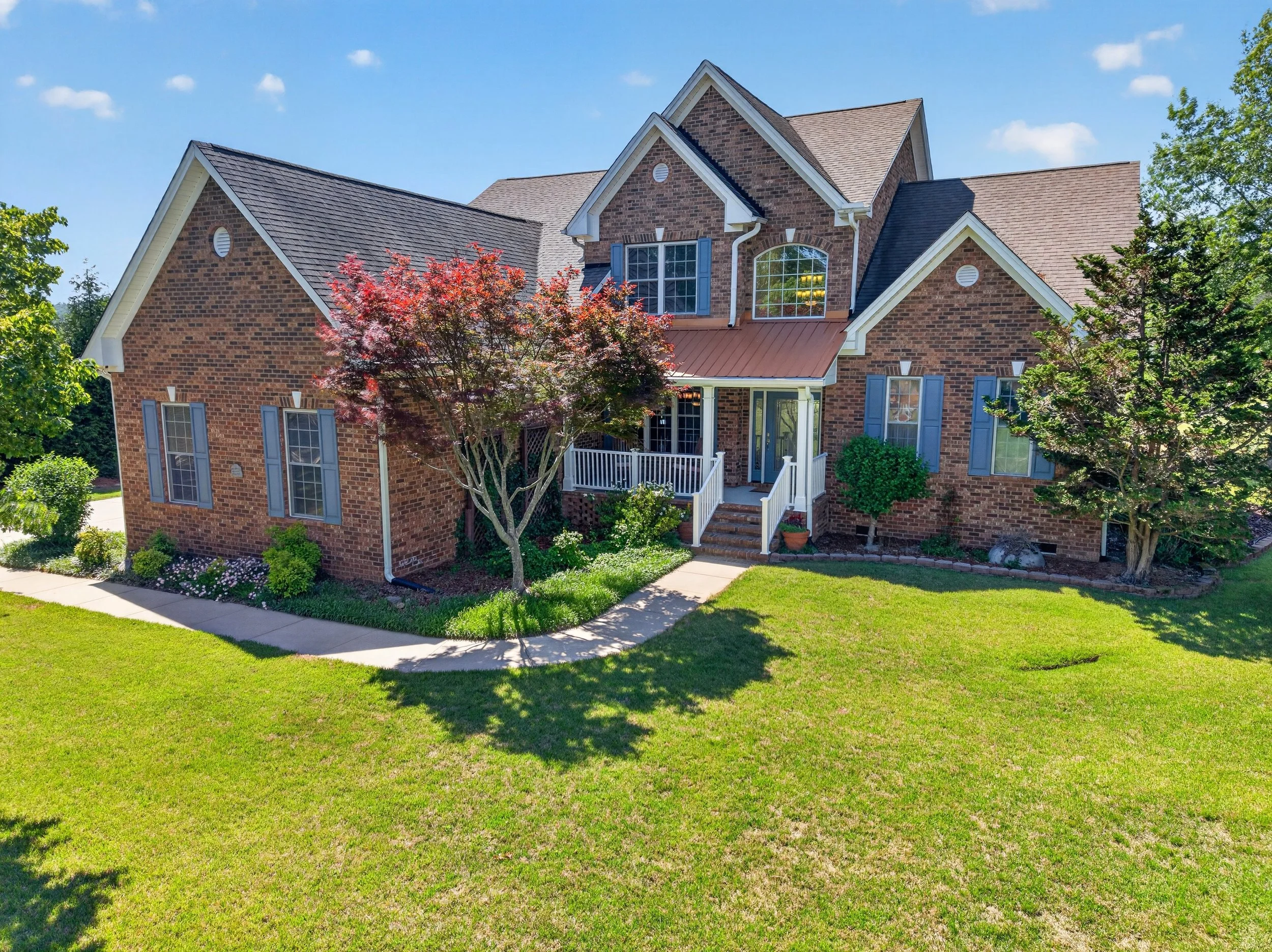 A large brick house with multiple gabled roofs, blue shutters, and a front porch with steps. There are trees and a green lawn in the front yard, under a bright blue sky with some clouds.