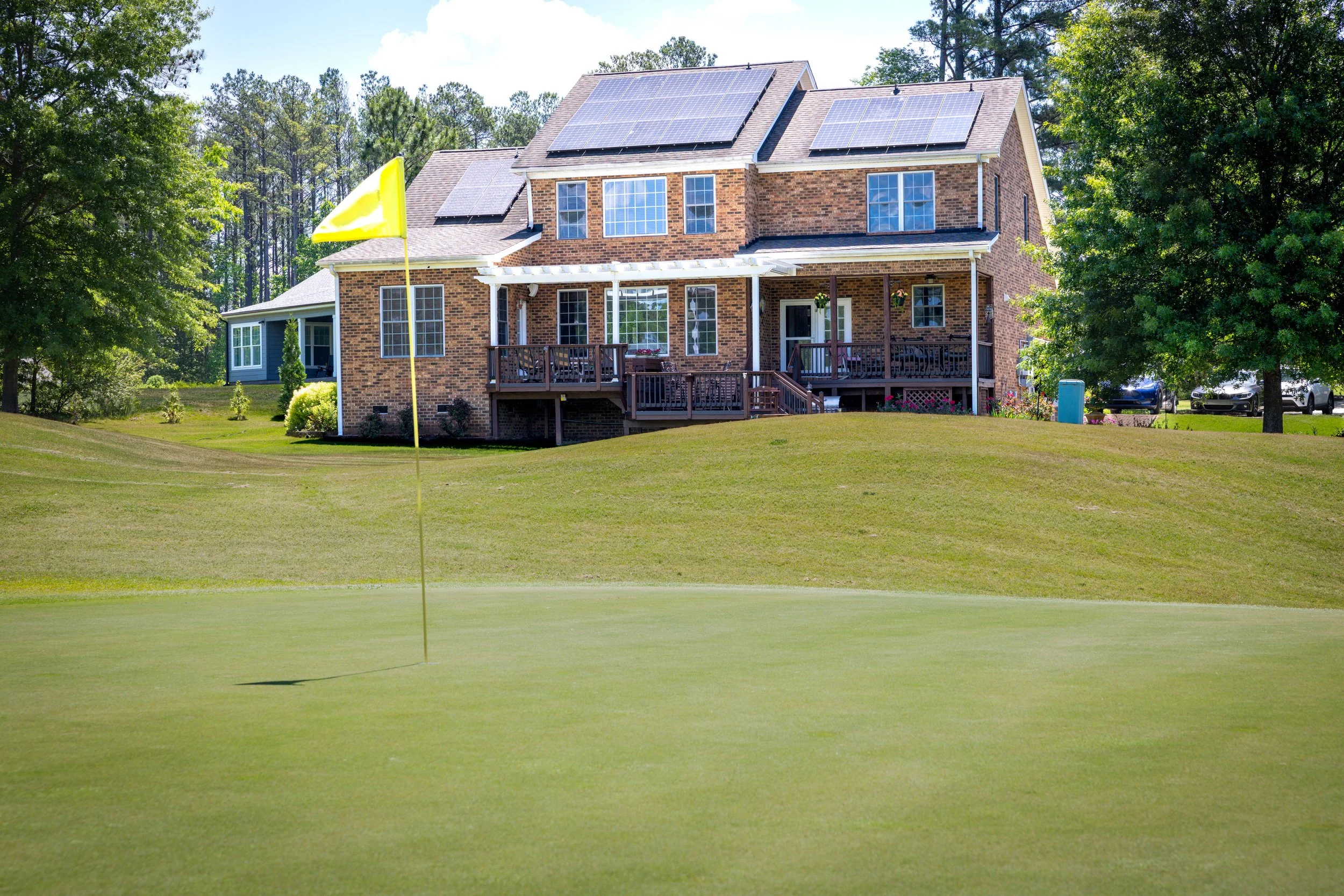 A large brick house with solar panels on the roof, surrounded by trees, and a deck area in the backyard. There is also a yellow golf flag in the foreground and a few parked cars in the background.