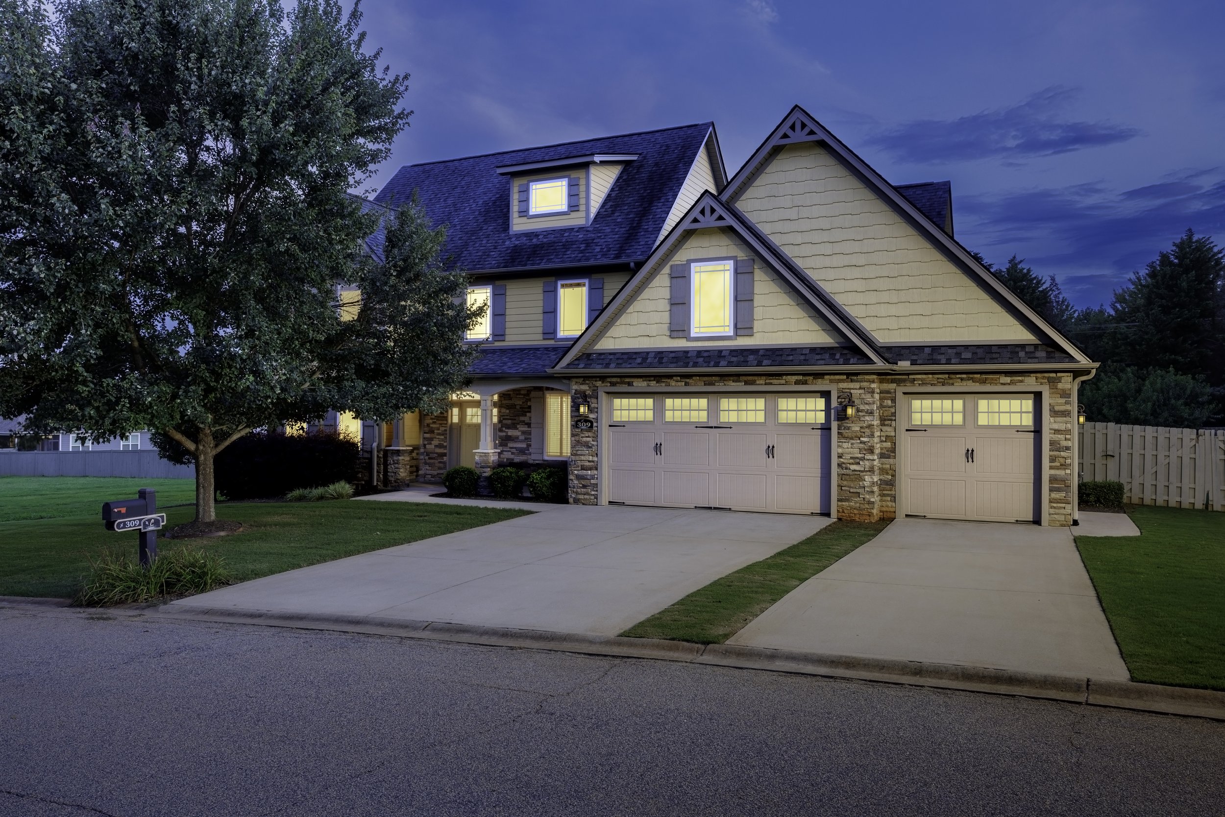 A two-story house with a yellow exterior and gray trim, illuminated from inside during the evening. The house features a garage with a double door, stone accents on the lower wall, and a small porch with white columns. There is a tree with full green leaves in the front yard, a mailbox, and a well-maintained lawn with a concrete pathway leading to the front door. The sky is partly cloudy with shades of blue.