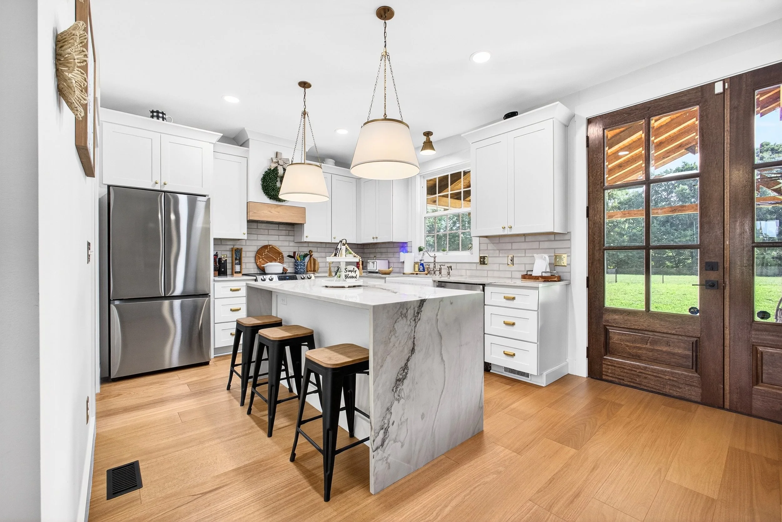 Modern white kitchen with a marble island, stainless steel refrigerator, and wooden stools. Large window and a glass door with wooden frames lead outside. Pendant lights hang over the island, and various kitchen items are on the countertops. Light wo