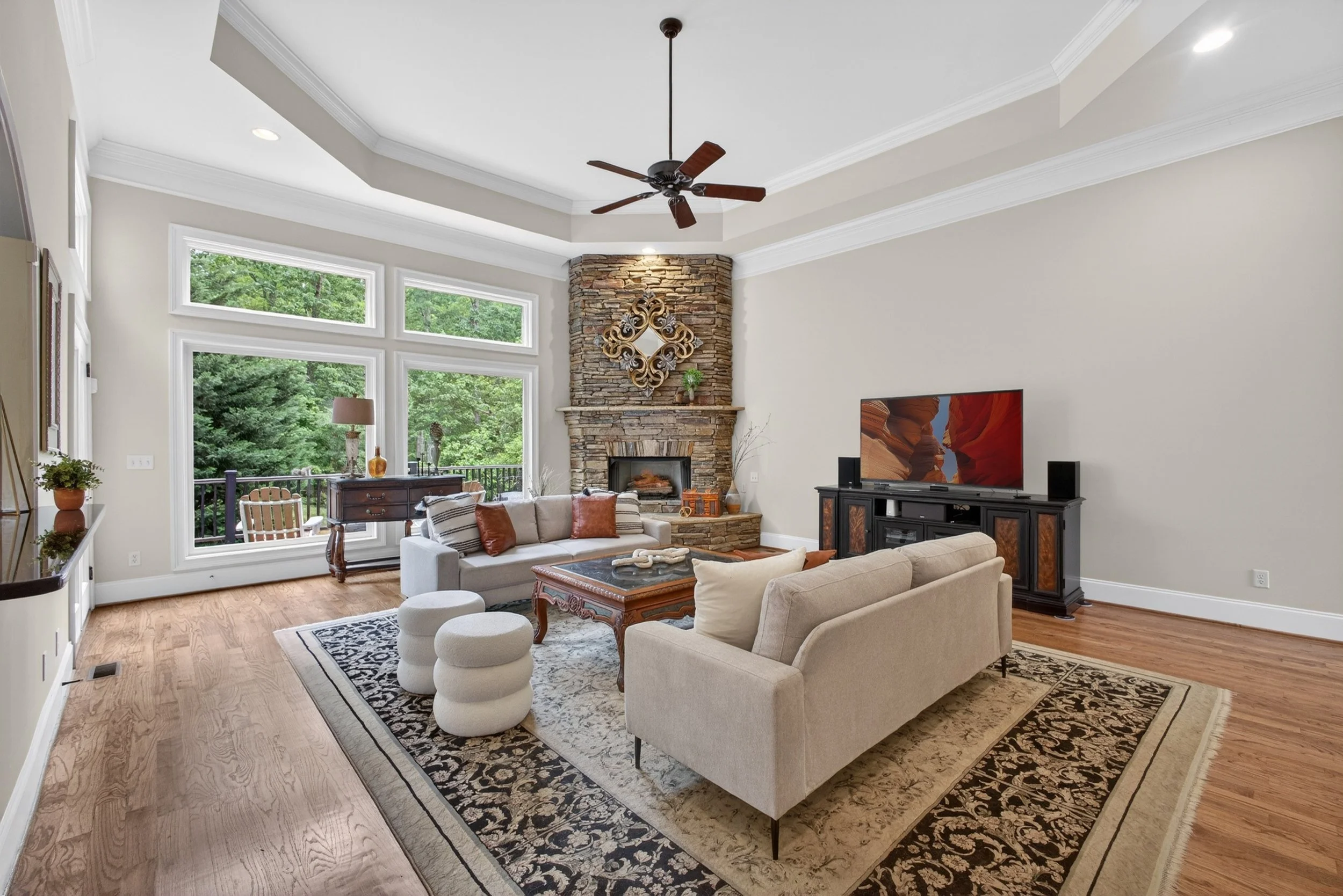 Living room with large windows showing green trees, a stone fireplace with a decorative mirror, a ceiling fan, a television, and beige furniture on a patterned rug.