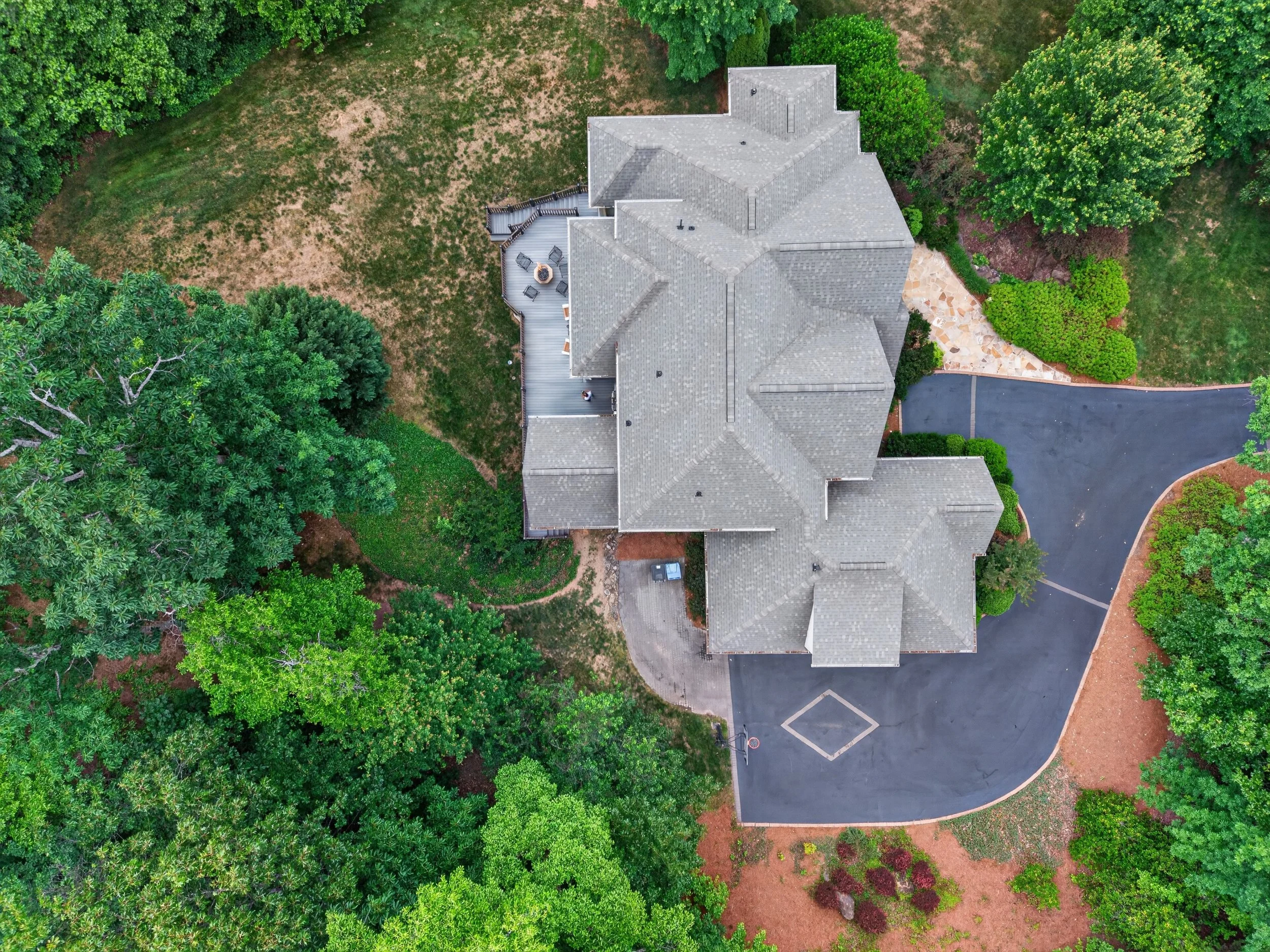 An aerial view of a house with a gray roof surrounded by trees, a backyard with a deck, and a parking lot with a basketball hoop.
