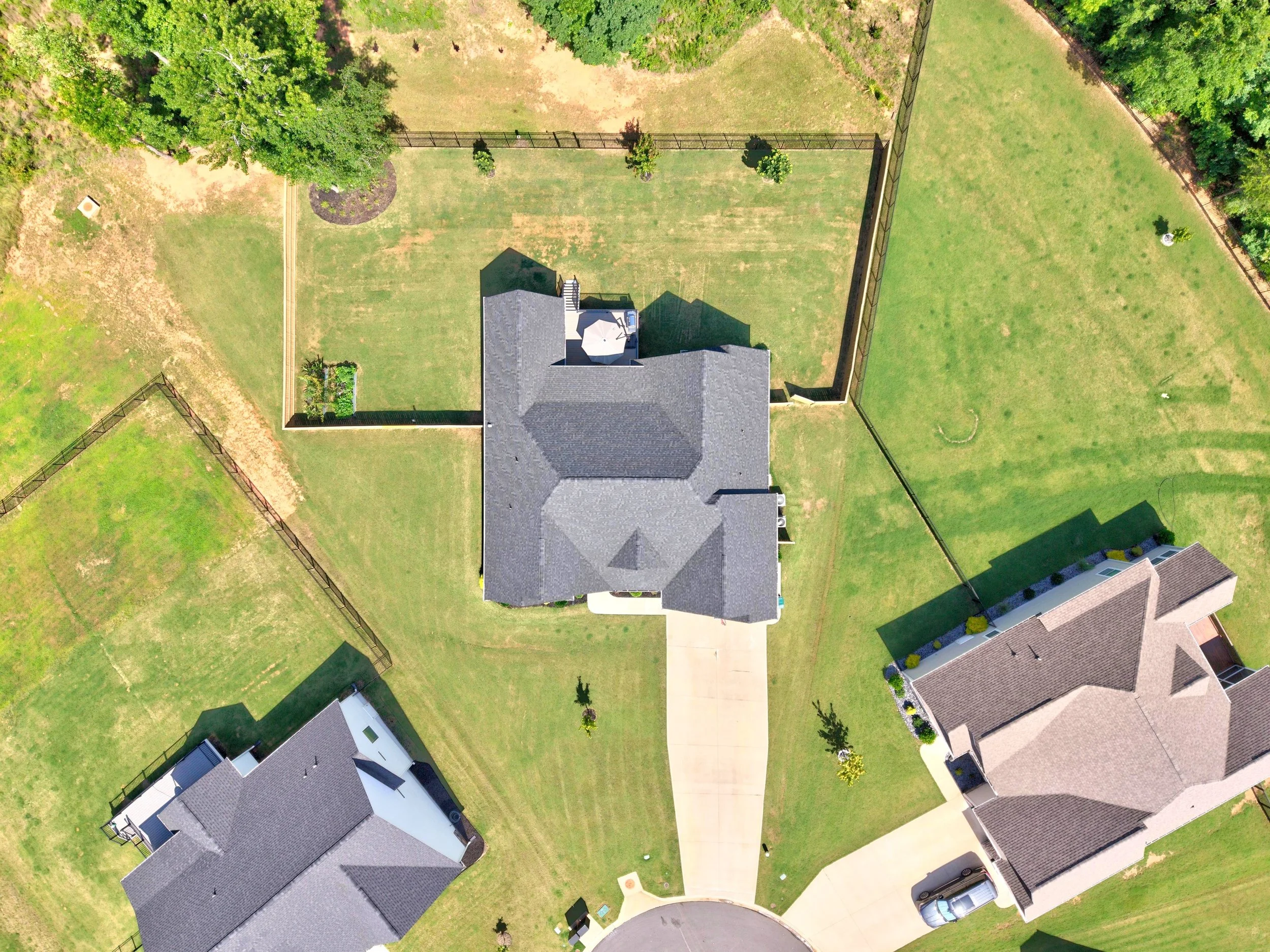 An aerial view of a backyard with green grass, a house with a gray roof, a patio with a table and umbrella, surrounded by a black fence, and some trees and neighboring houses.
