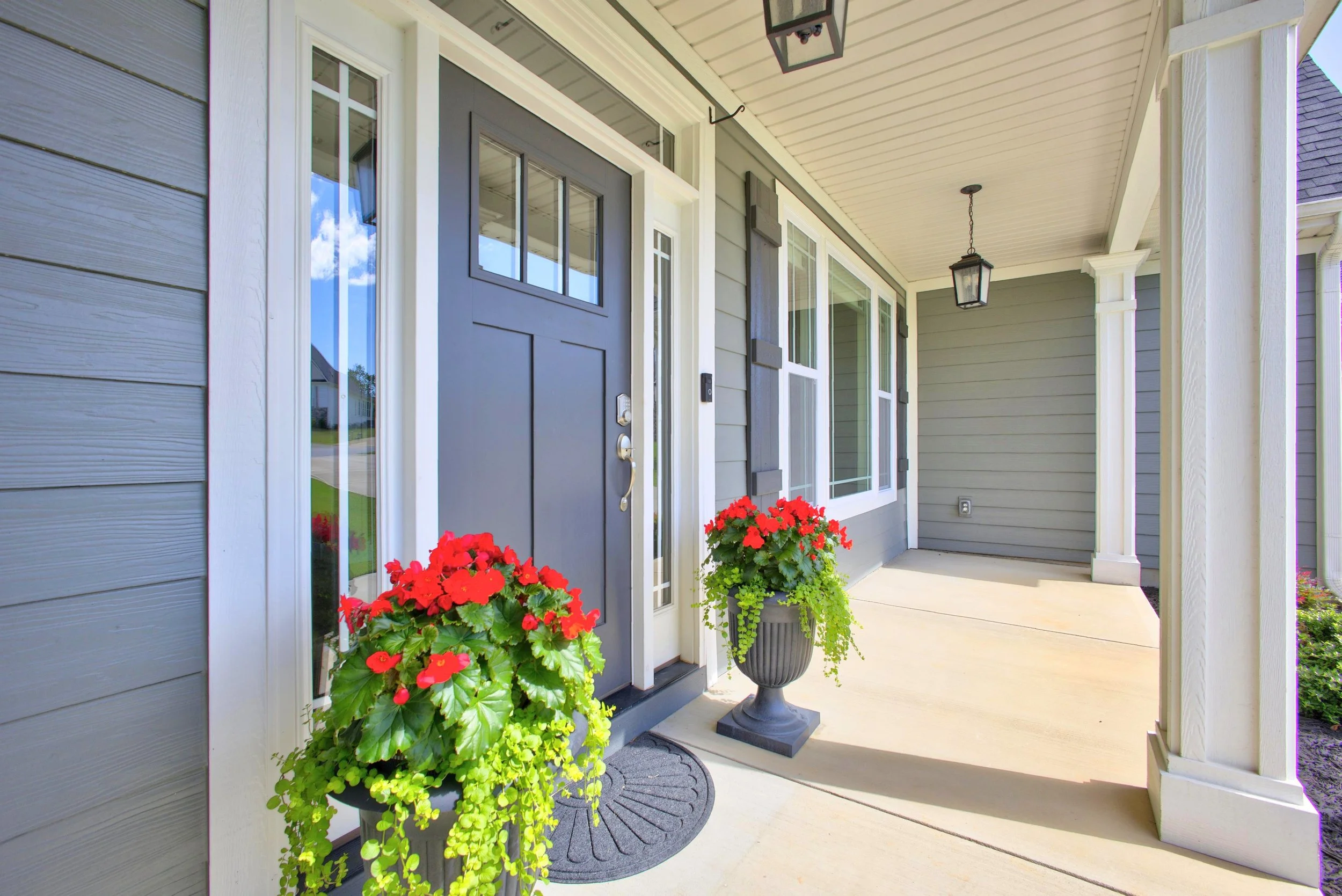 Front porch of a house with gray siding, a dark gray door, and two hanging lanterns. There are two large potted plants with red and green foliage on each side of the door, a gray doormat, and a concrete walkway. The porch has white trim and columns.