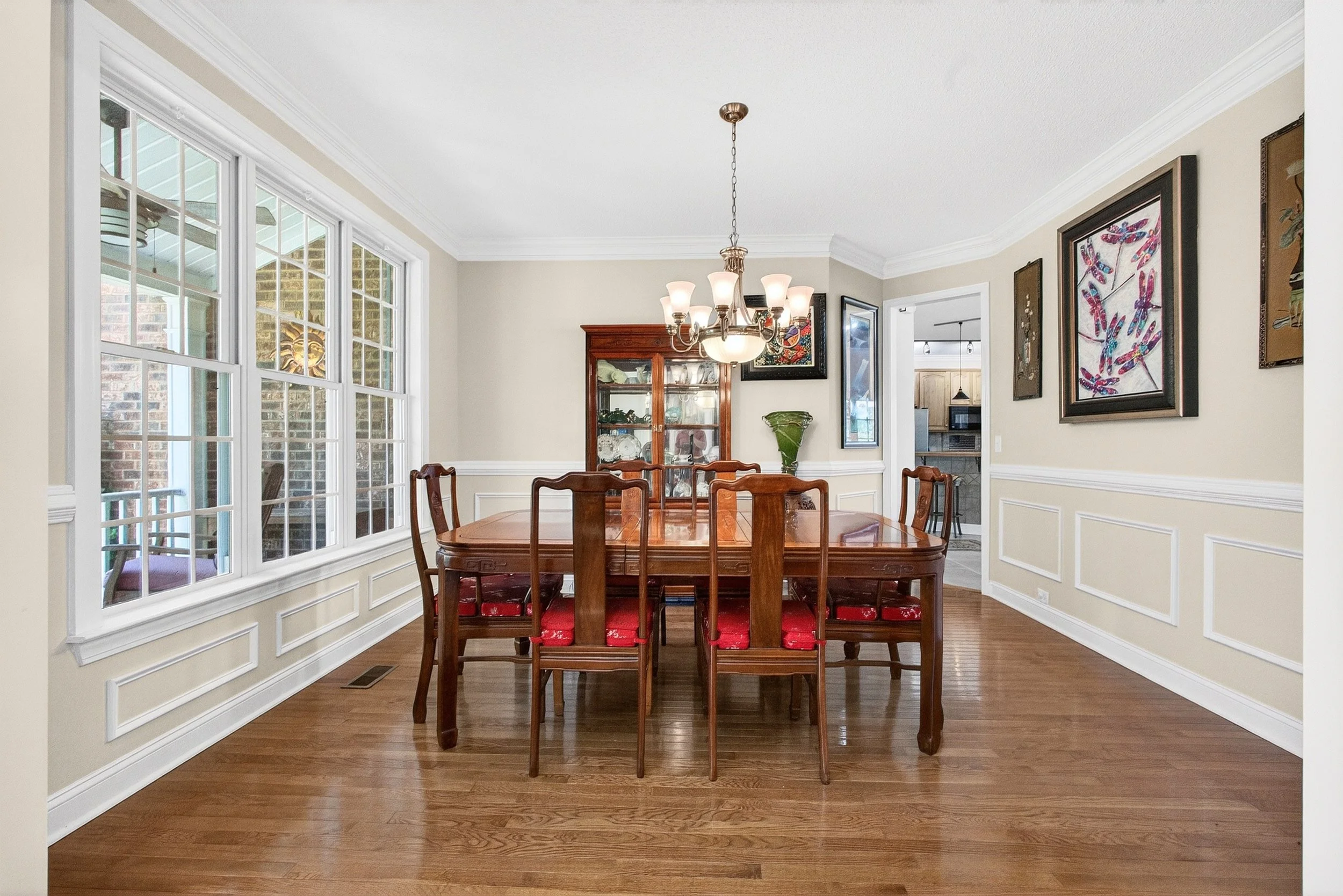 Dining room with large window, wooden table with six chairs, chandelier, corner display cabinet, wall art, and hardwood floors.
