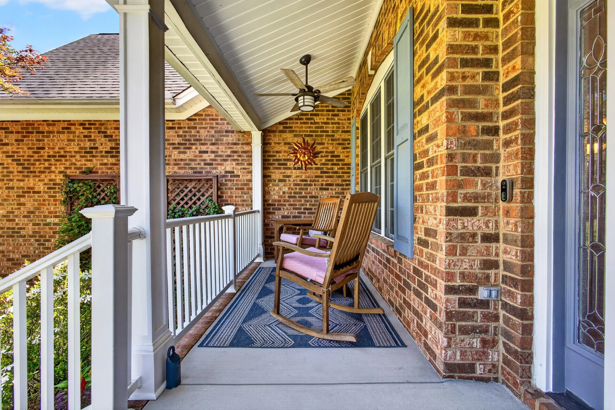 Covered front porch with brick walls, white railing, two wooden rocking chairs with pink cushions, ceiling fan, sun sculpture, ventilation grille, exterior door with decorative glass, and a gardening water bottle on the porch floor.
