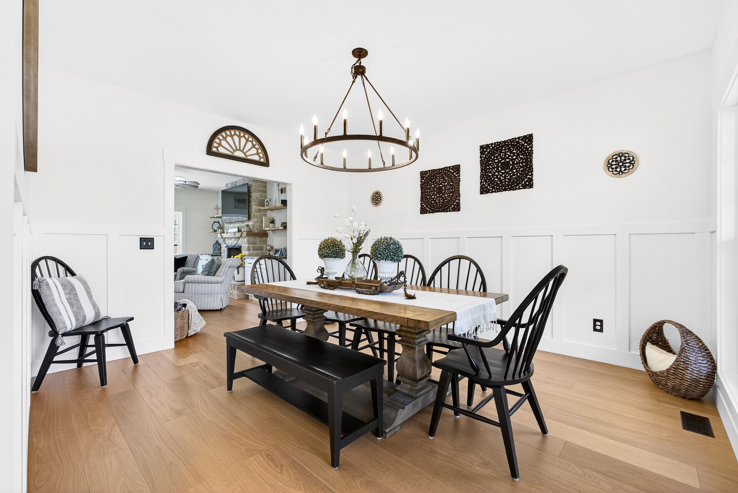 A dining room with a wooden dining table, black chairs, and a modern chandelier. The wall decor includes black and brown decorative panels and small circular wall hangings. A hallway leads to a living area with a brick fireplace and a TV.