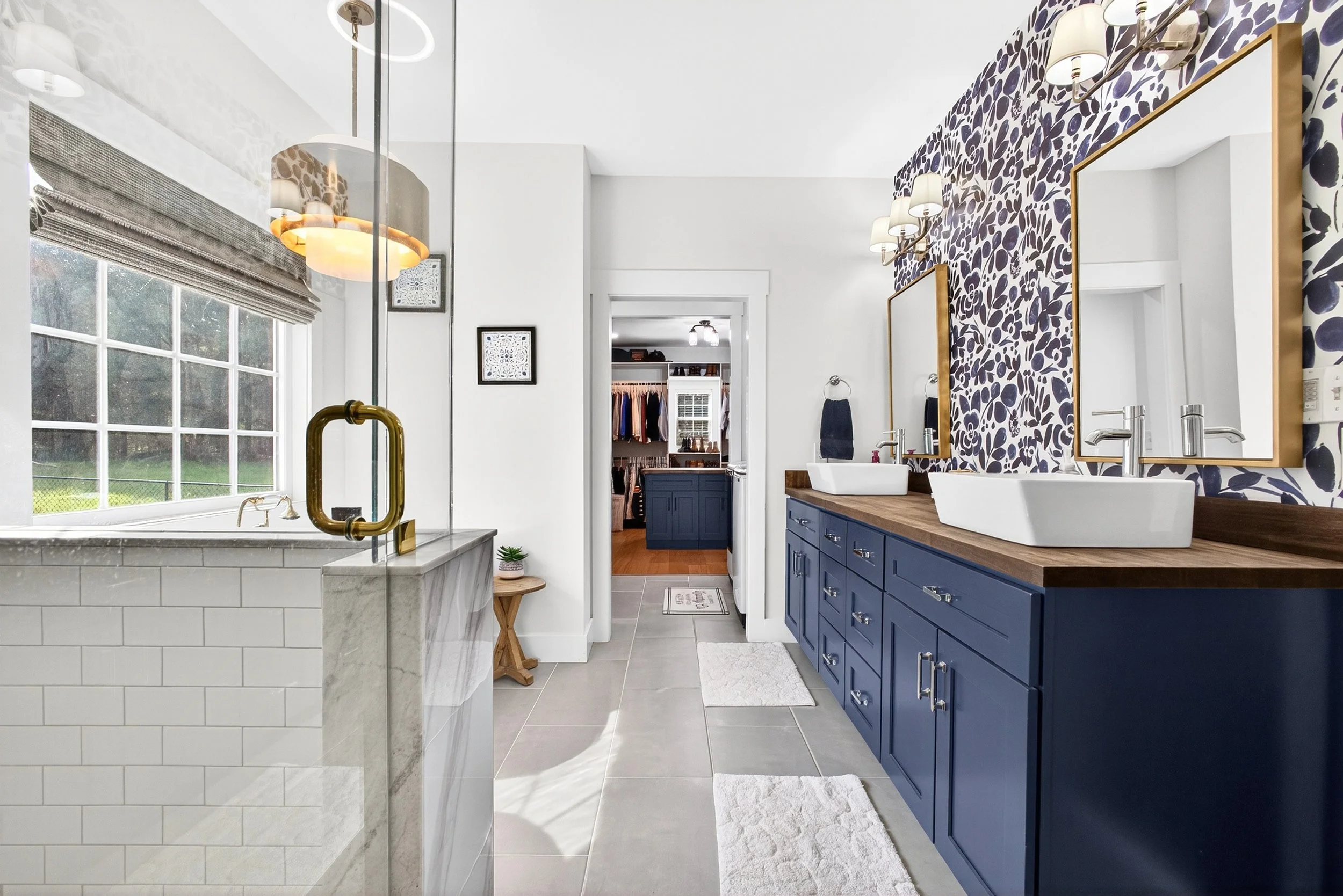 Bathroom with large window, navy blue cabinets, white double vessel sinks, patterned wallpaper, and a view of a walk-in closet in the background.