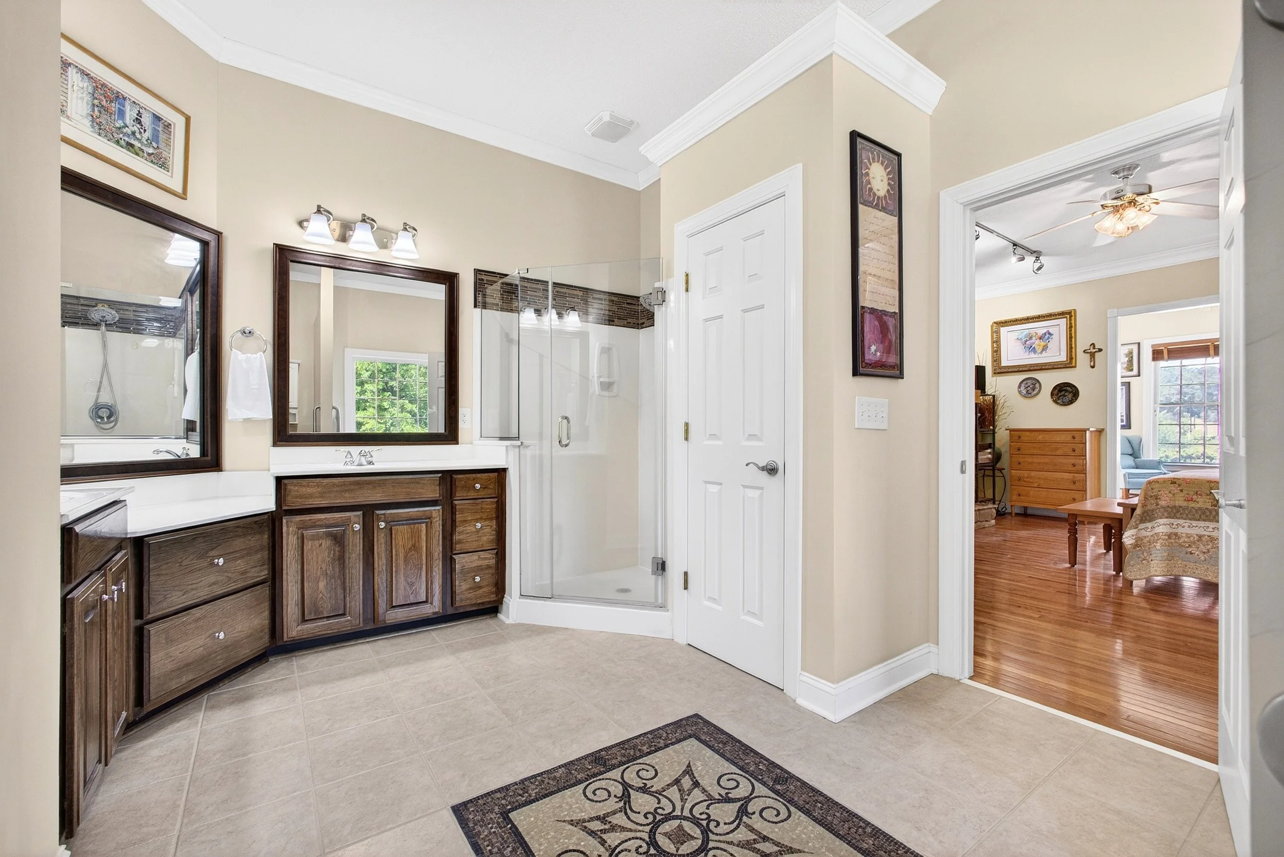 A bathroom with a vanity mirror, dark wood cabinets, a glass shower, and a towel ring. Adjacent to the bathroom are two doorways leading to a living space with hardwood floors and furniture.