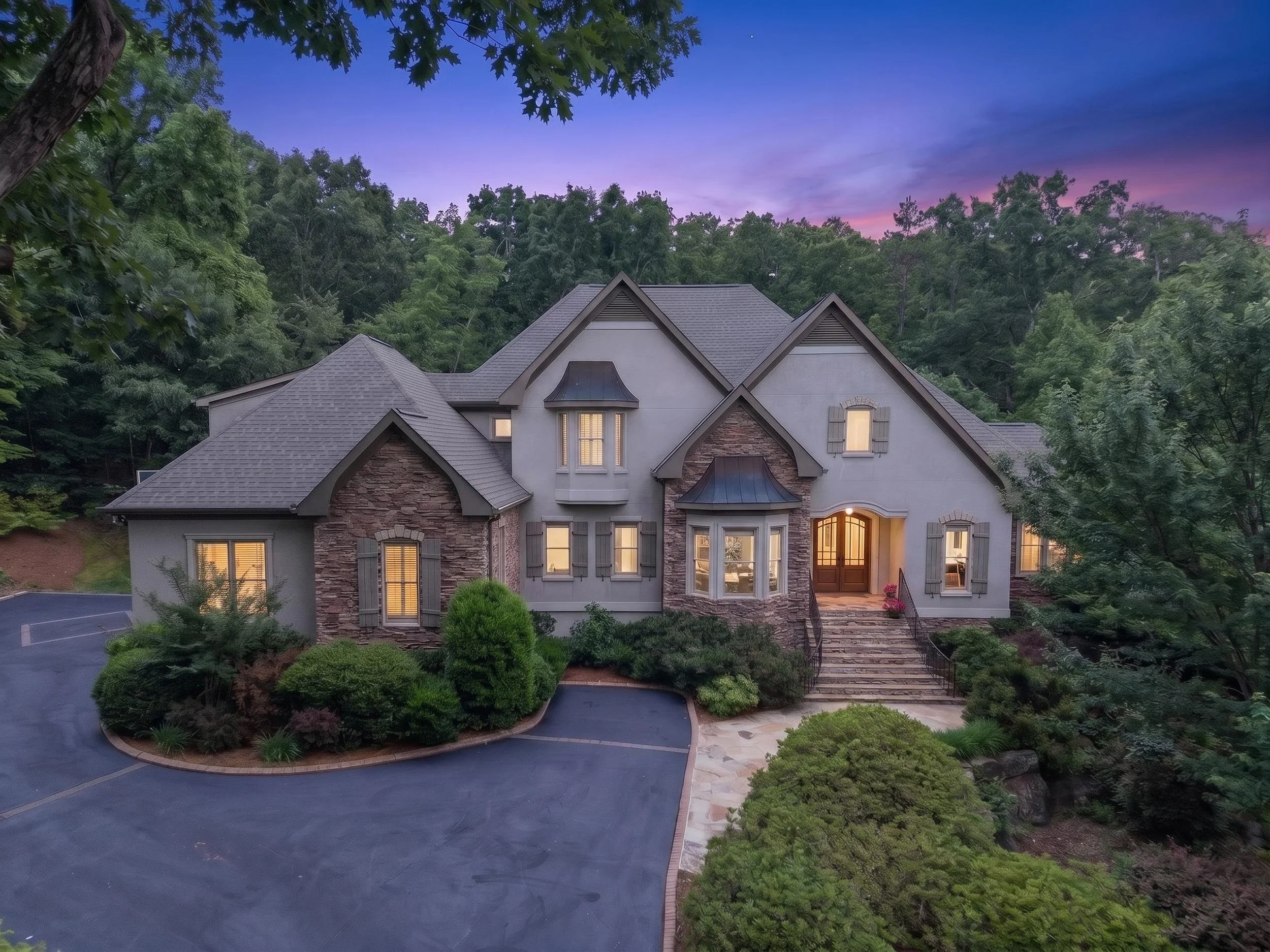 A large house with a detailed roof and a porch, surrounded by a landscaped yard with bushes and trees, under a twilight sky with clouds and a hint of purple and pink hues.
