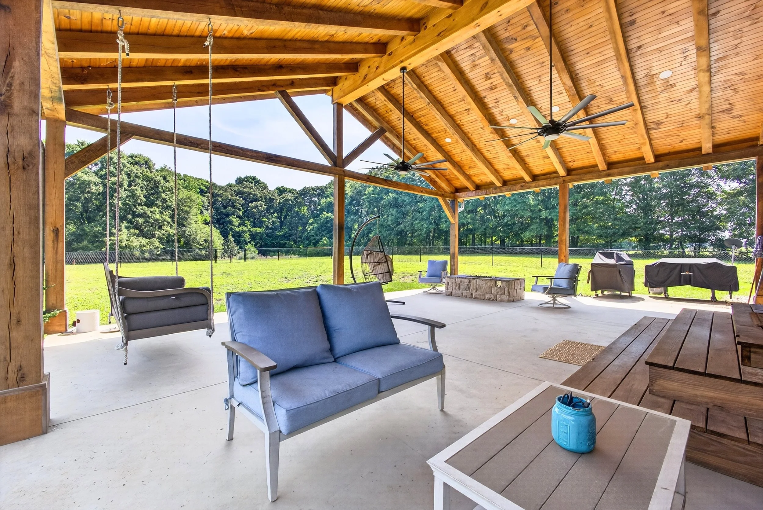 Covered outdoor patio with ceiling fans, outdoor furniture including a blue loveseat, hanging swing chair, and a picnic table. There is a grassy yard with trees and a fence in the background.