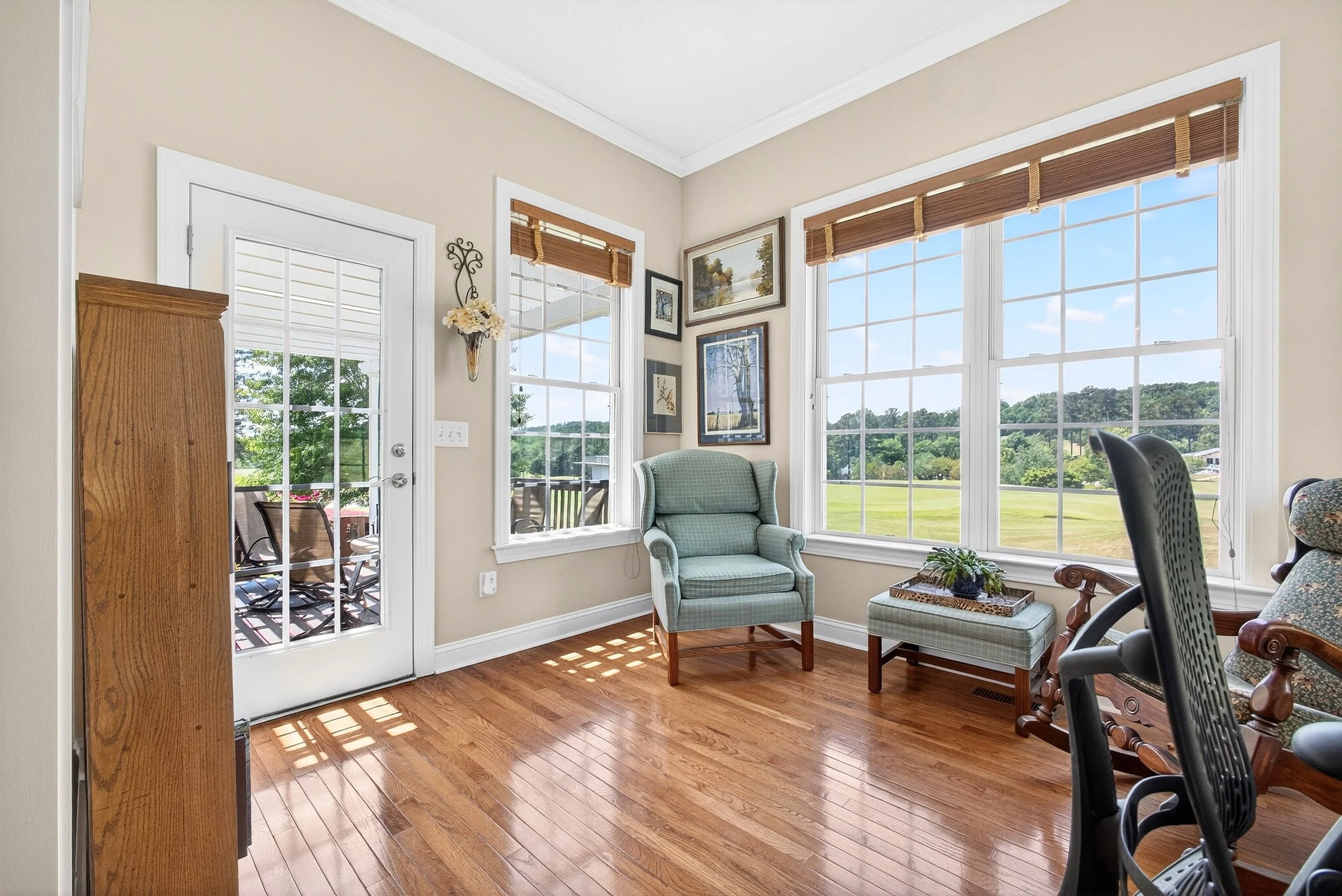 Living room with large windows, wooden floor, armchair, framed artwork on wall, glass door leading outside, and a glimpse of outdoor patio and yard.