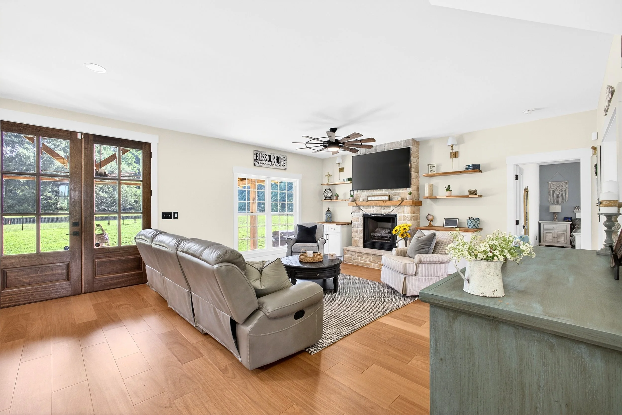 Living room with beige sofa, armchair, fireplace, wooden shelves, flat-screen TV, ceiling fan, large windows, and decorative items.