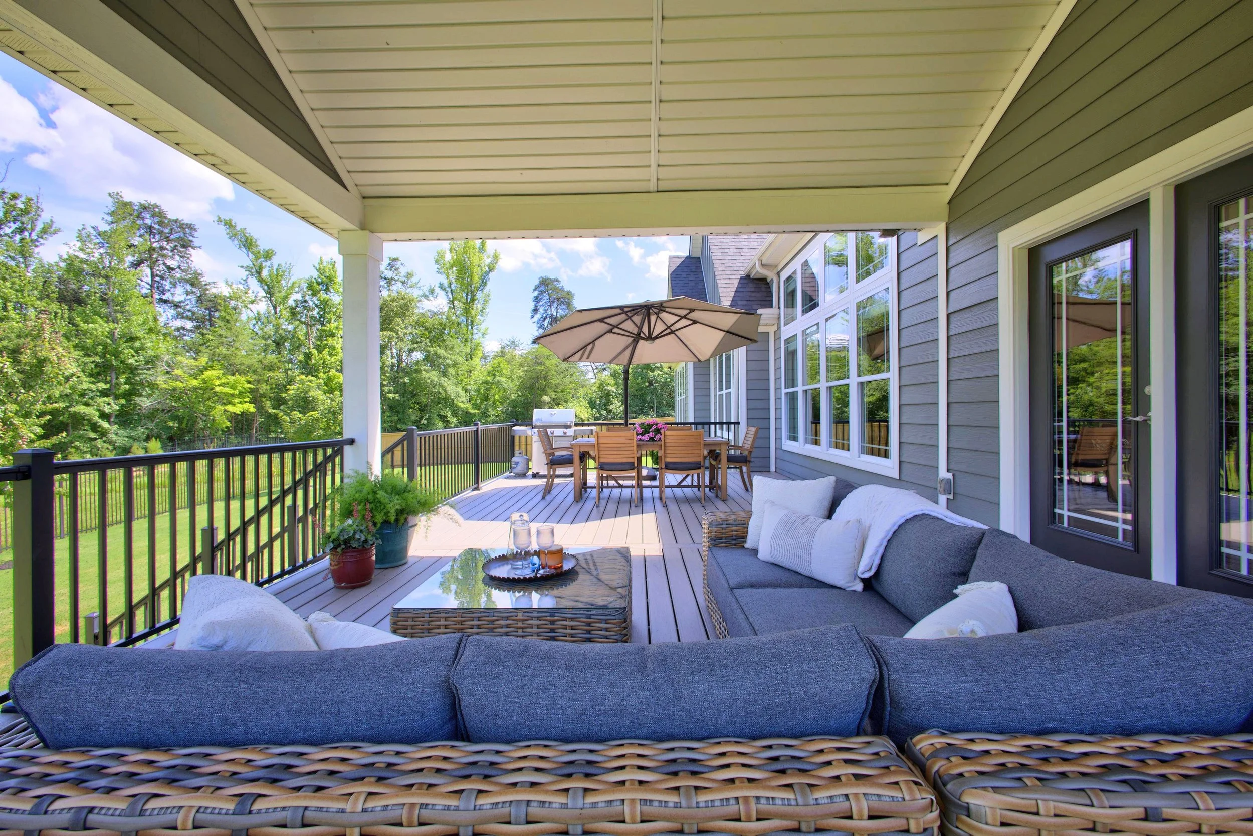 Outdoor patio with wicker furniture, cushions, a coffee table with beverages, and an umbrella. Surrounding green trees and a fence.