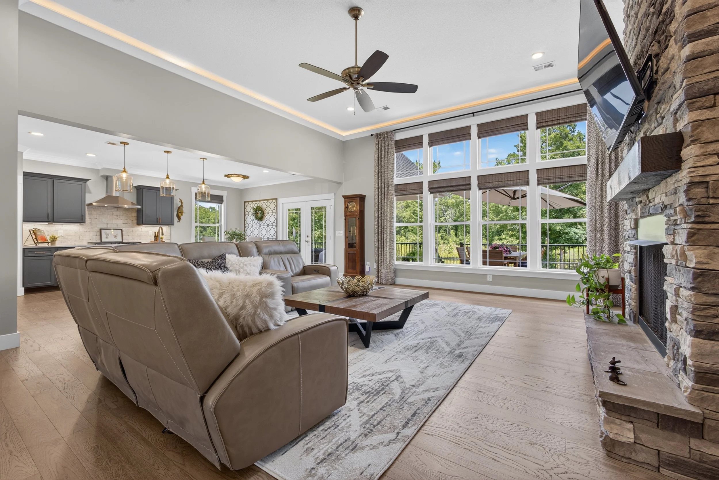 Bright living room with large bay window, stone fireplace, ceiling fan, gray kitchen cabinets, and modern decor.