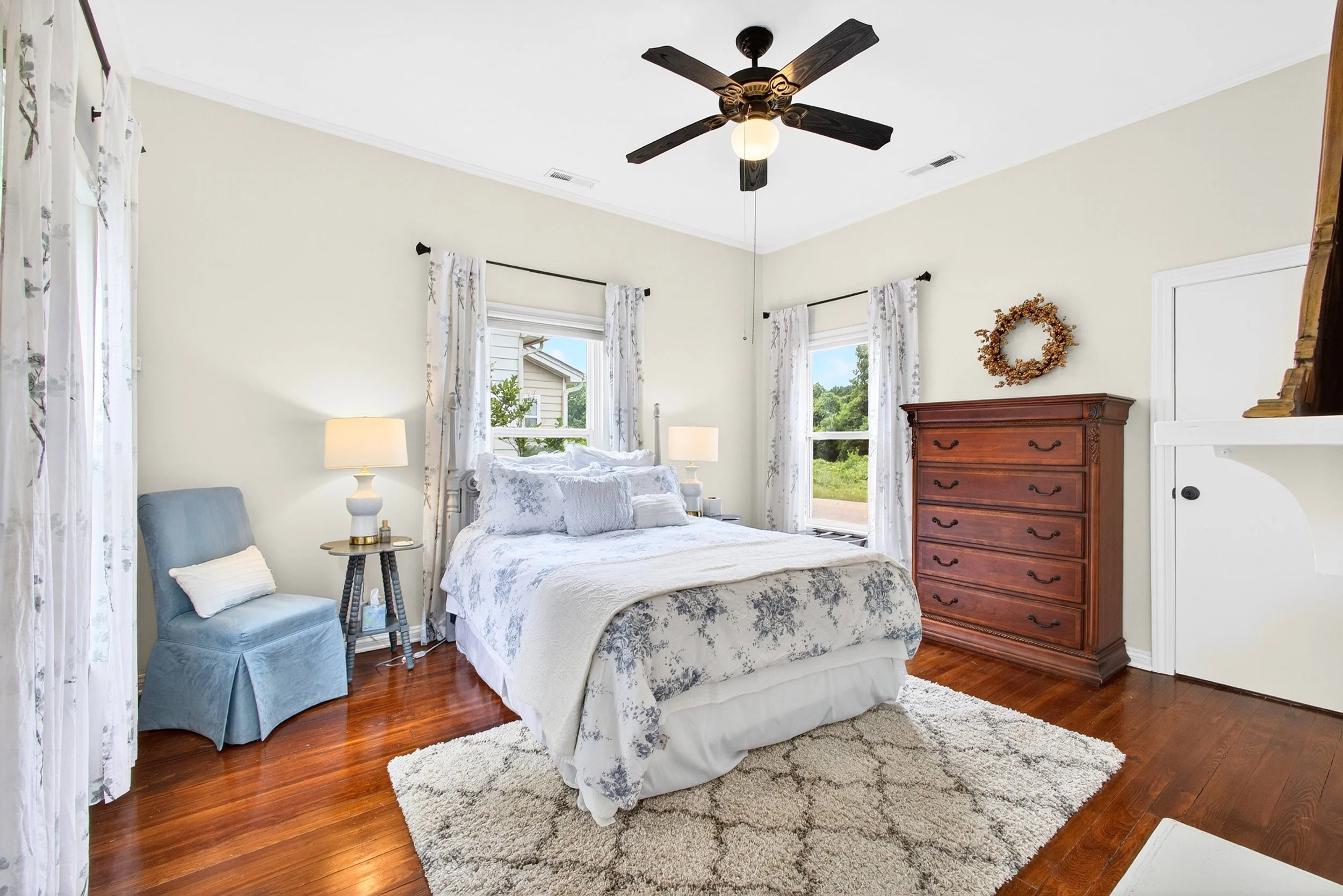 A bedroom with a wooden floor, cream walls, and large windows with white curtains. There is a bed with white and gray floral bedding, a wooden dresser, a blue armchair with a pillow, and a white shaggy rug. A ceiling fan with wooden blades hangs from
