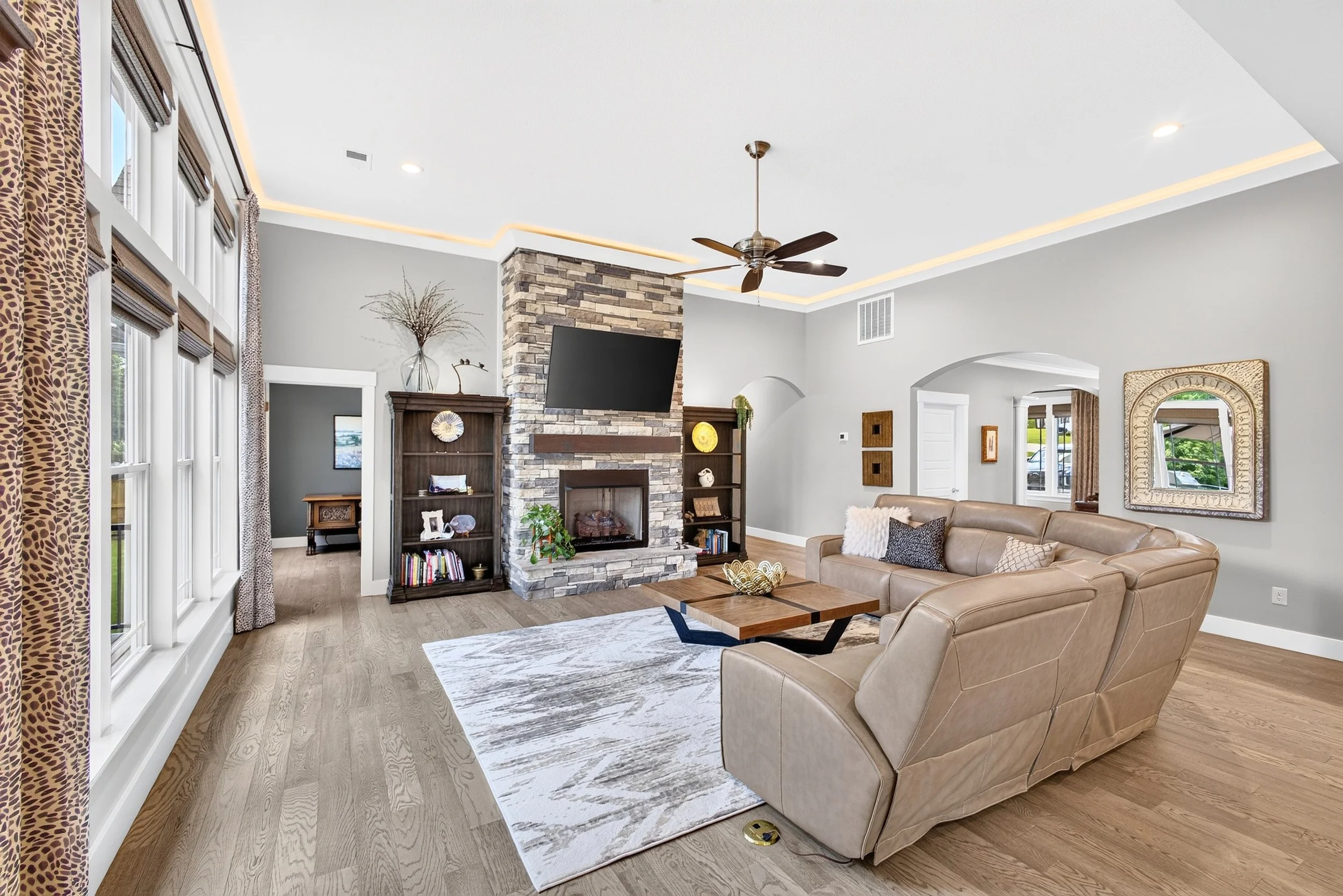 Living room with large window, beige sectional sofa, stone fireplace with a TV above it, and various decorative items on shelves and walls.