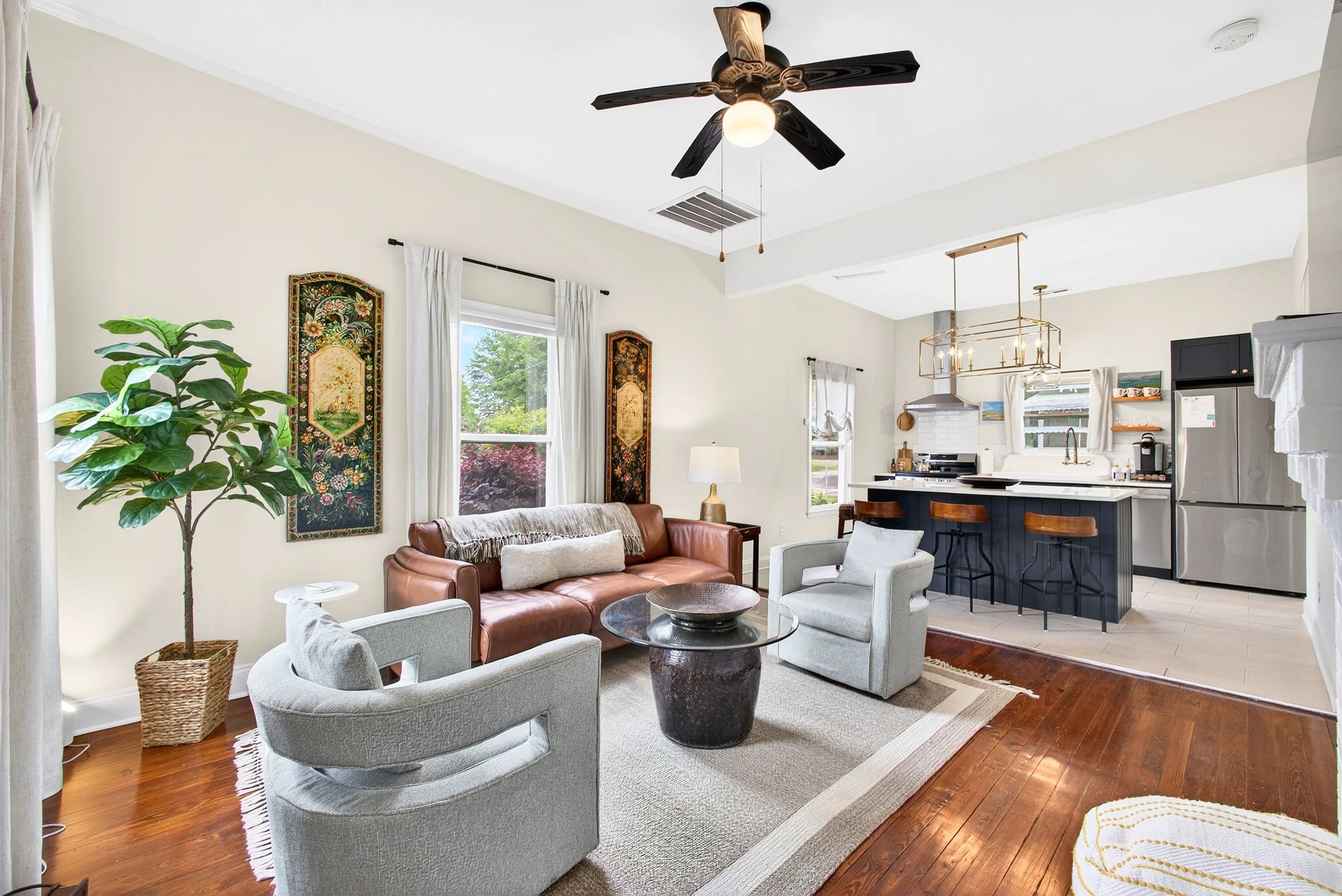Living room with a beige sofa, gray armchair, white curtains, a large potted plant, and wall decorations, open to a kitchen with black island, white cabinets, and stainless steel appliances.