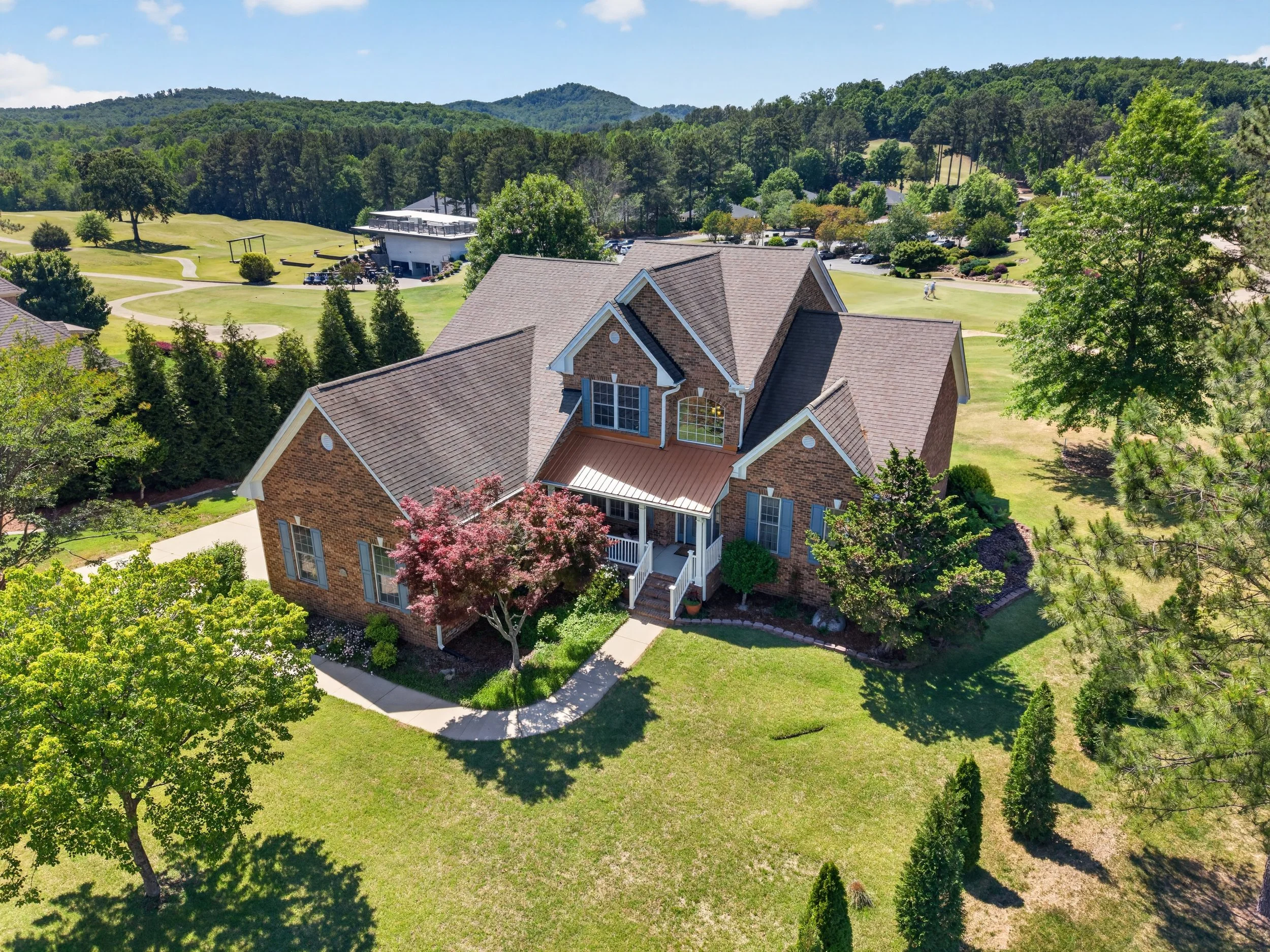 Aerial view of a suburban house with a brick exterior, surrounded by green trees, a manicured lawn, and a walking path, with a golf course and lush forested hills in the background on a clear sunny day.