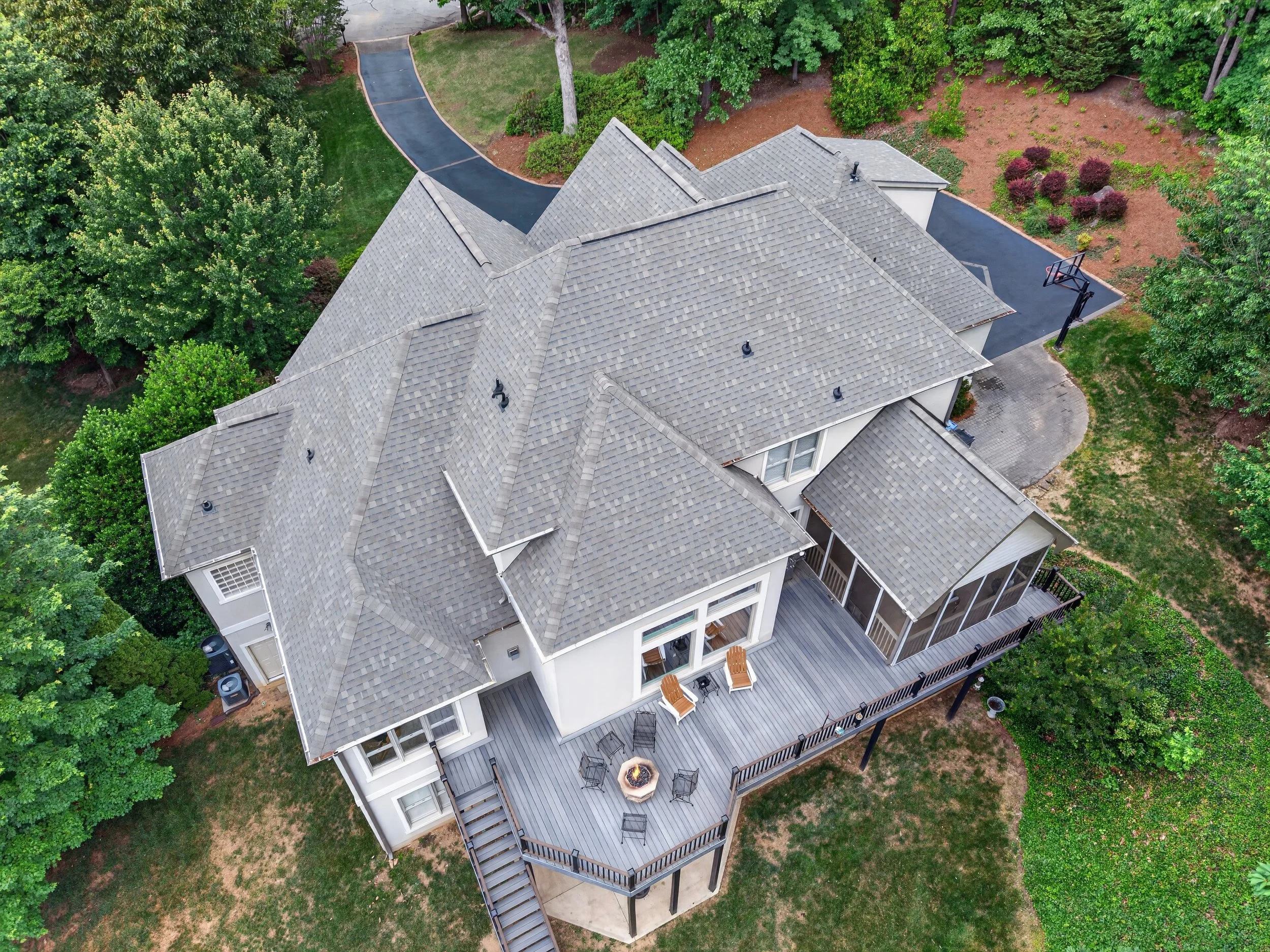 Aerial view of a house with a gray shingle roof, black asphalt driveway, and a wooden deck with outdoor furniture, surrounded by green trees and landscaped yard.